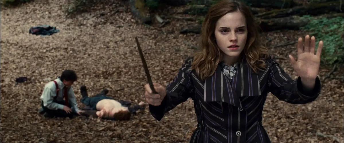 Hermione Granger (Emma Watson) stands over Harry Potter (Daniel Radcliffe) and Ron Weasley (Rupert Grint) on the ground, wand raised and one palm out, casting a shield.