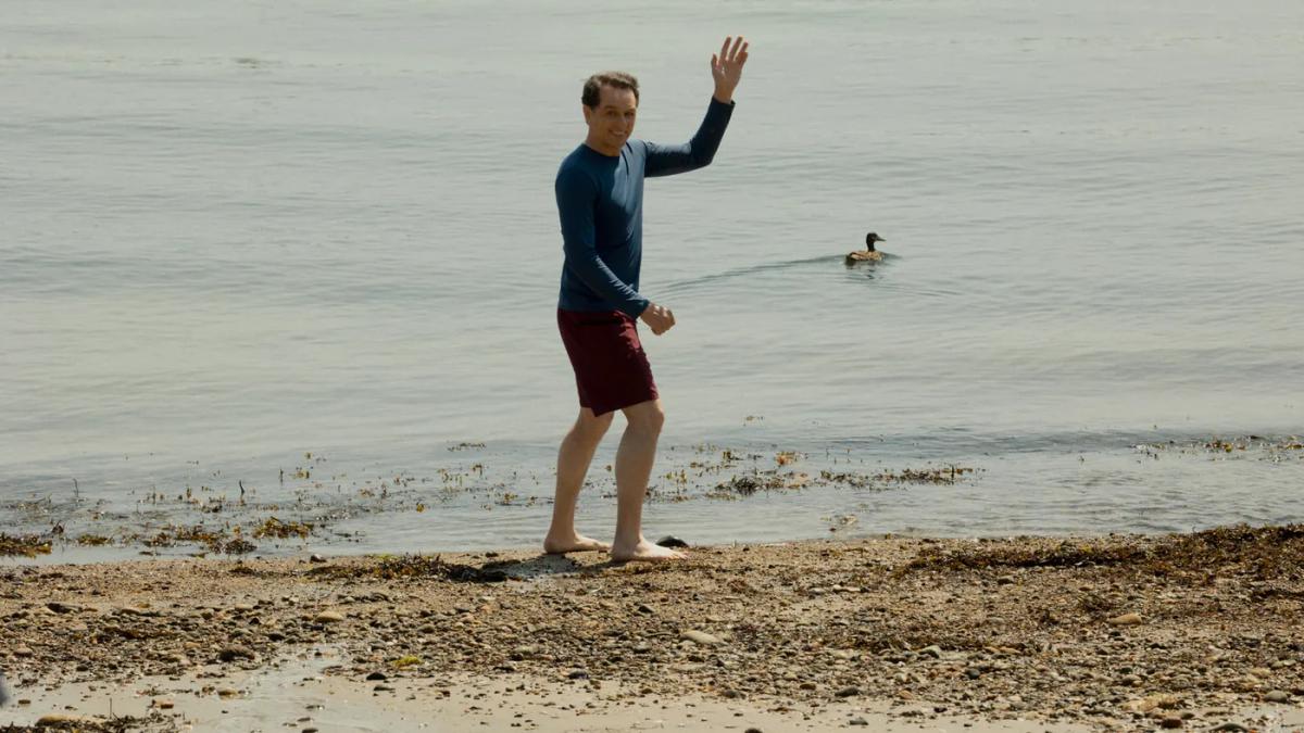 Matthew Rhys as Tom Loftis stands barefoot at the shoreline in a rash vest and swim shorts, waving to someone off camera, with a duck paddling behind him.