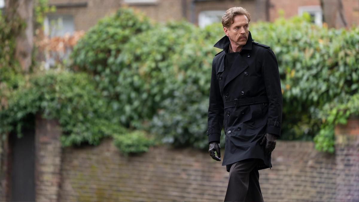 Damian Lewis as the Caller walks along a suburban London pavement in a dark trench coat and leather gloves.