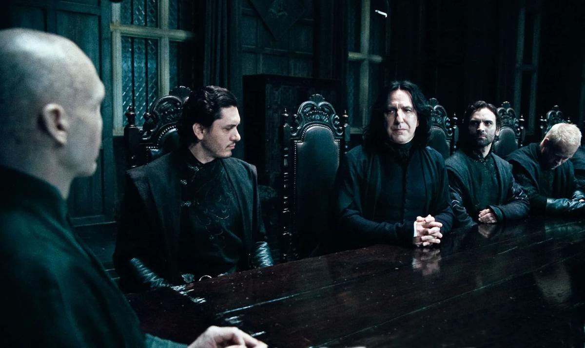 Lord Voldemort (Ralph Fiennes) sits in the foreground at the head of a long dark table while Severus Snape (Alan Rickman) and other Death Eaters sit in a row facing him.