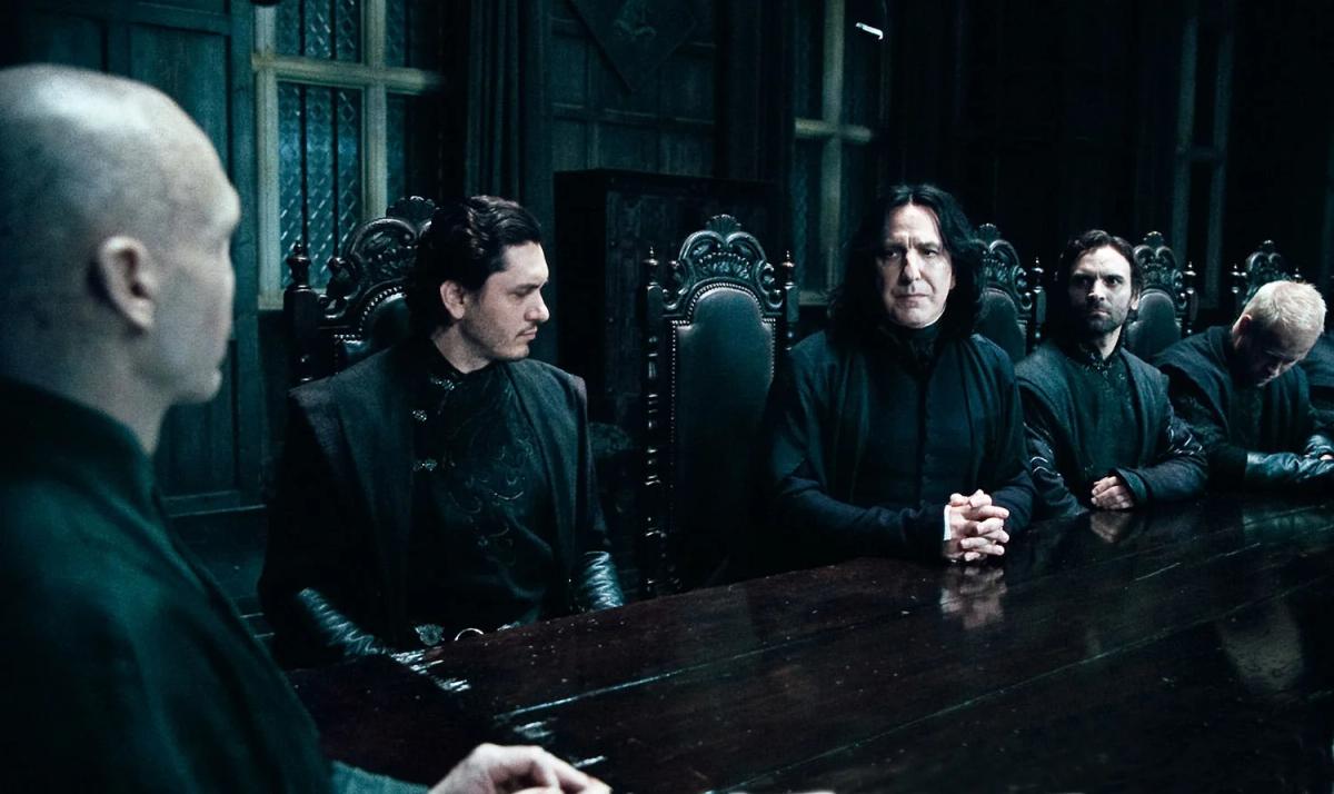 Lord Voldemort (Ralph Fiennes) sits in the foreground at the head of a long dark table while Severus Snape (Alan Rickman) and other Death Eaters sit in a row facing him.