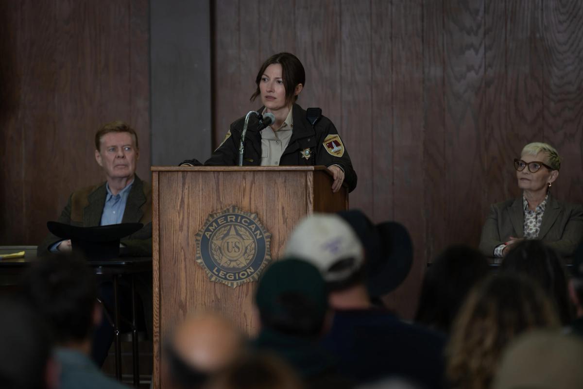Kelly Macdonald as Sheriff Kerry Kane speaking into a microphone at a wooden podium bearing an American Legion seal, addressing a seated crowd.