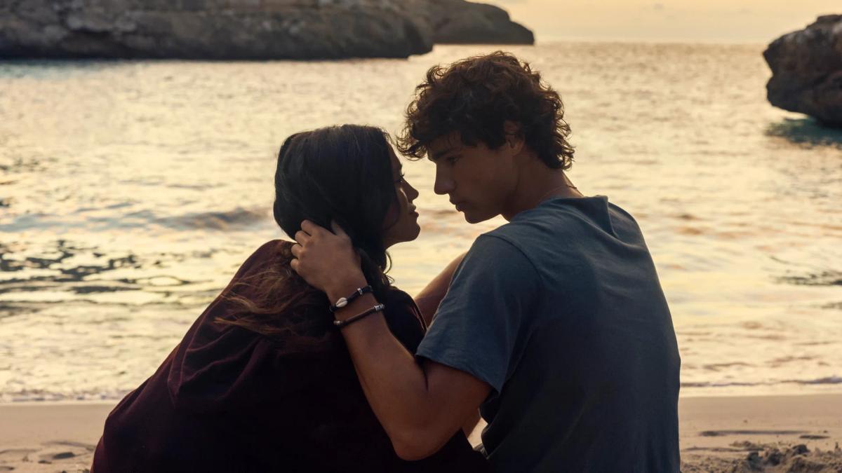 Maia Reficco as Oriah "Ry" Pera wrapped in a blanket on the beach at sunset, with Fernando Lindez as Julián García holding her.