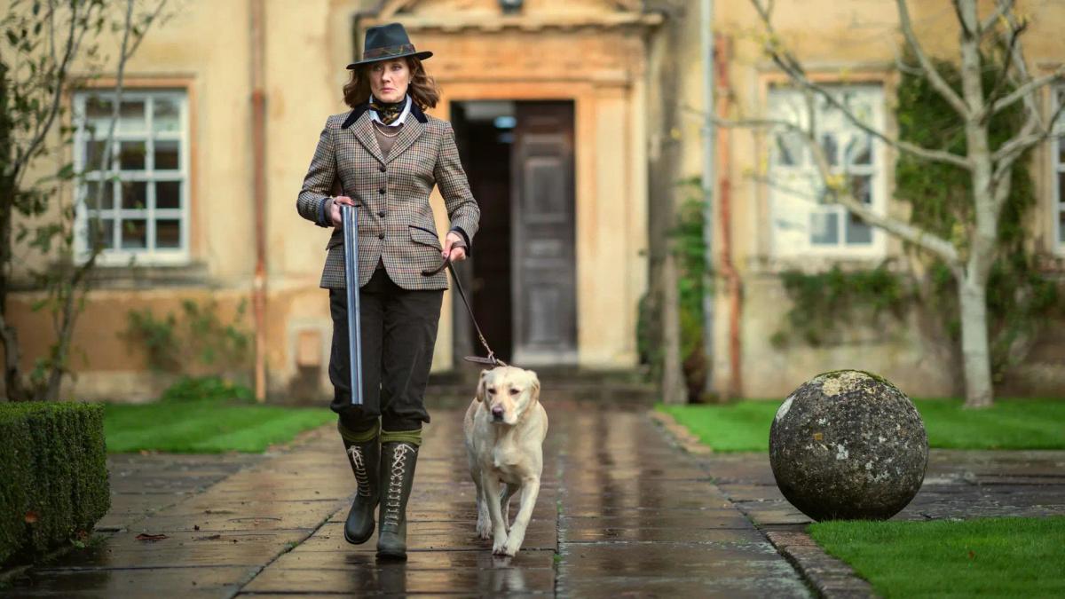 Joely Richardson as Lady Sabrina Horniman walking a labrador down a wet path at Halstead, carrying a broken shotgun.