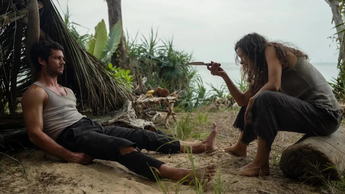 Rachel McAdams as Linda Liddle crouches holding a knife upright while Dylan O'Brien as Bradley Preston sits barefoot against a palm-frond shelter, looking away.