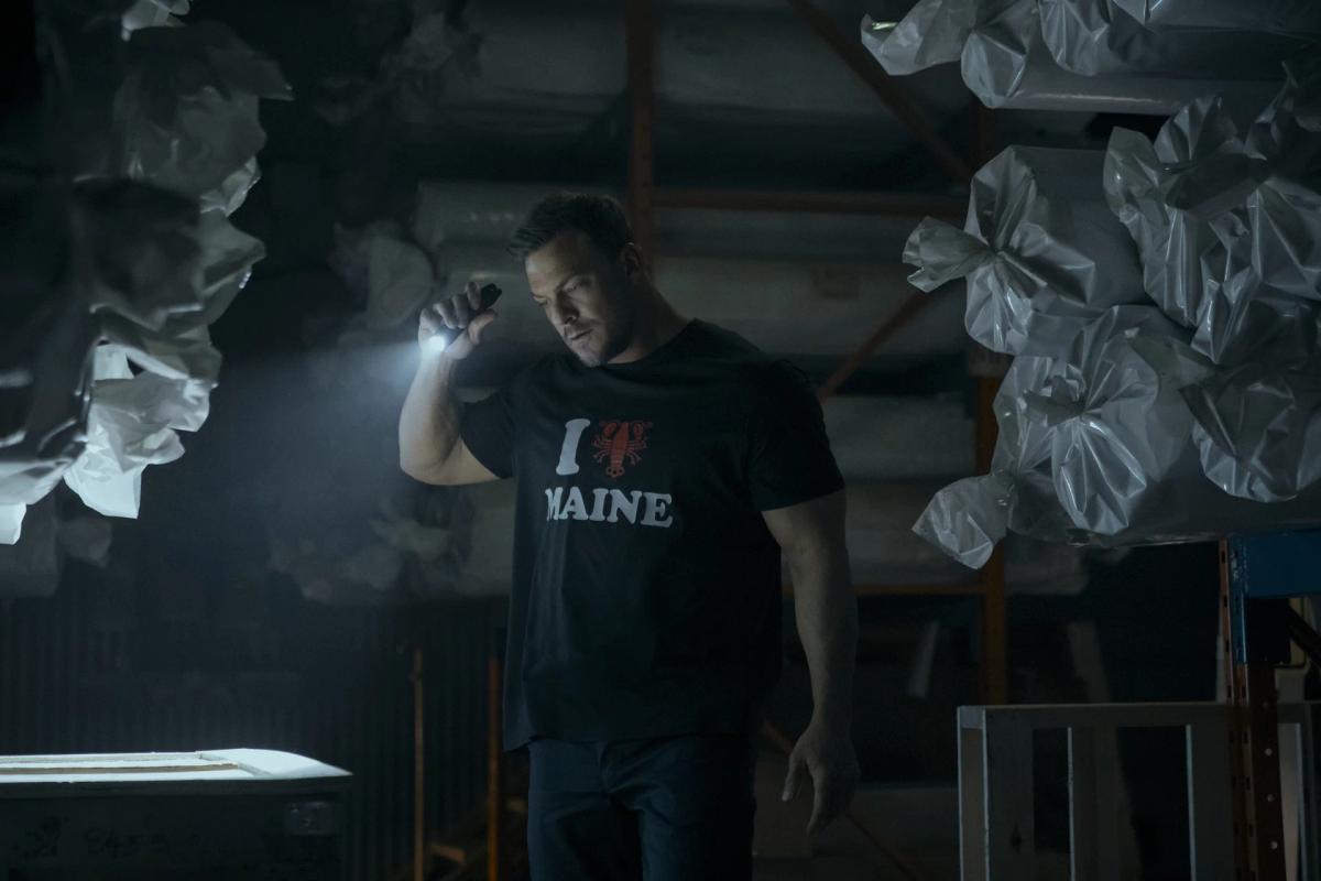 Alan Ritchson as Jack Reacher searches a dark warehouse by flashlight, wearing a black I Love Maine T-shirt with a red lobster in place of the heart, between stacks of plastic-wrapped rolls.