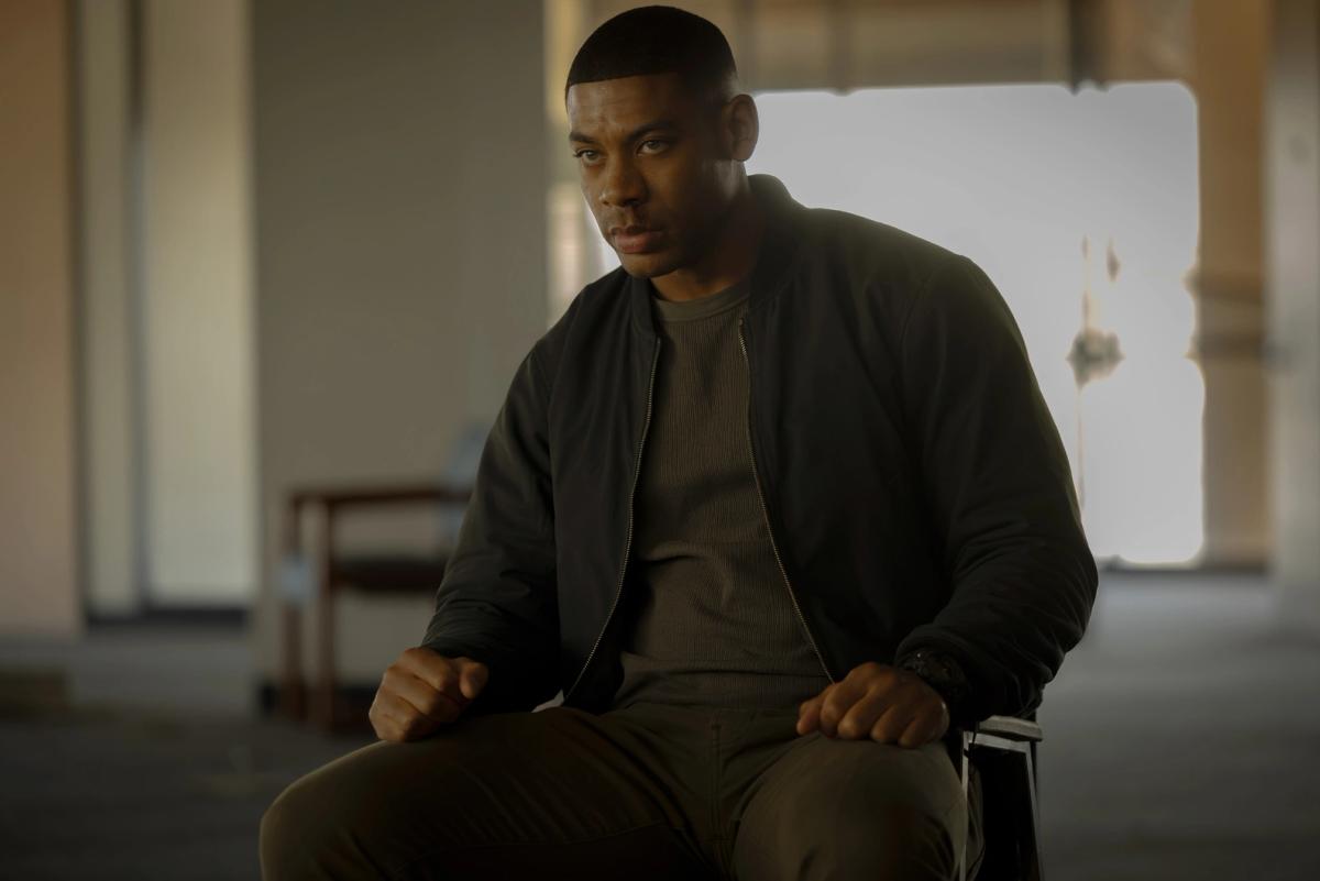 Aaron Pierre as John Stewart sitting on a chair in a dim, near-empty room, hands resting on his knees, looking off to one side.