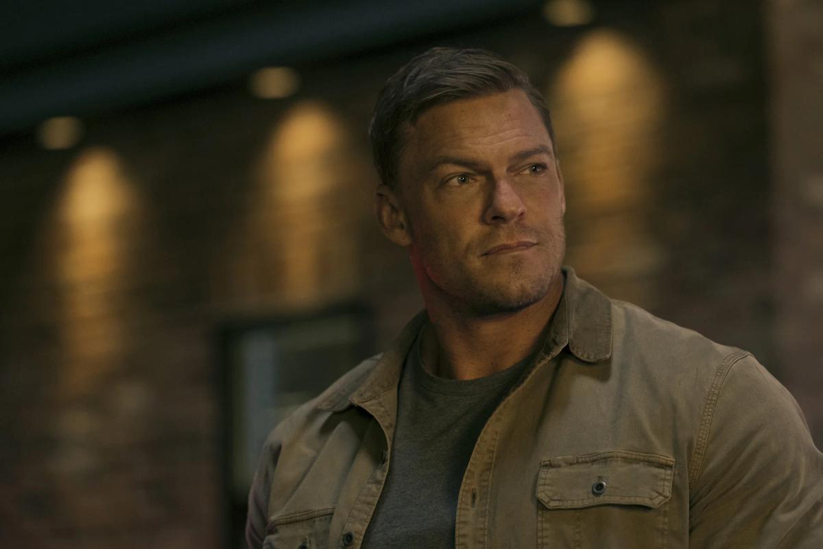 Close-up of Alan Ritchson as Jack Reacher in a canvas jacket over a green T-shirt, lit warm from the side, looking off-frame with the beginnings of a smile.