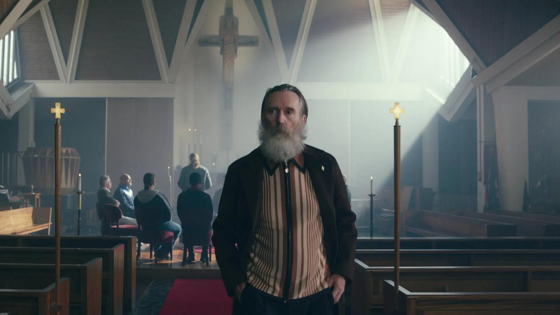Pearce Quigley as Gospel John standing in the aisle of his church, a crucifix behind him and a small congregation seated to one side.