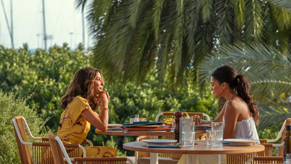 Eva Longoria as Isolde Pera and Maia Reficco as Oriah "Ry" Pera sitting opposite each other at an outdoor restaurant table.