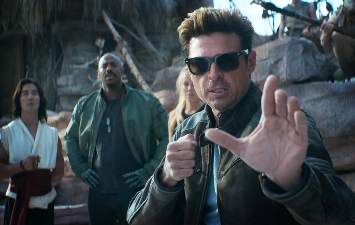 Karl Urban as Johnny Cage in sunglasses and a leather jacket, raising his hands in a fighting pose, with Mehcad Brooks as Jax and Jessica McNamee as Sonya Blade behind him.