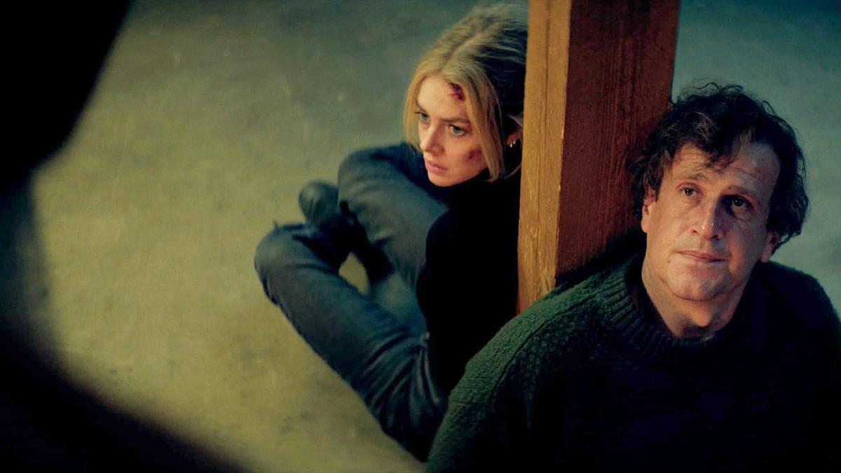Samara Weaving as Lisa crouching behind a post with a pistol while Jason Segel as Dan sits bloodied in the foreground.