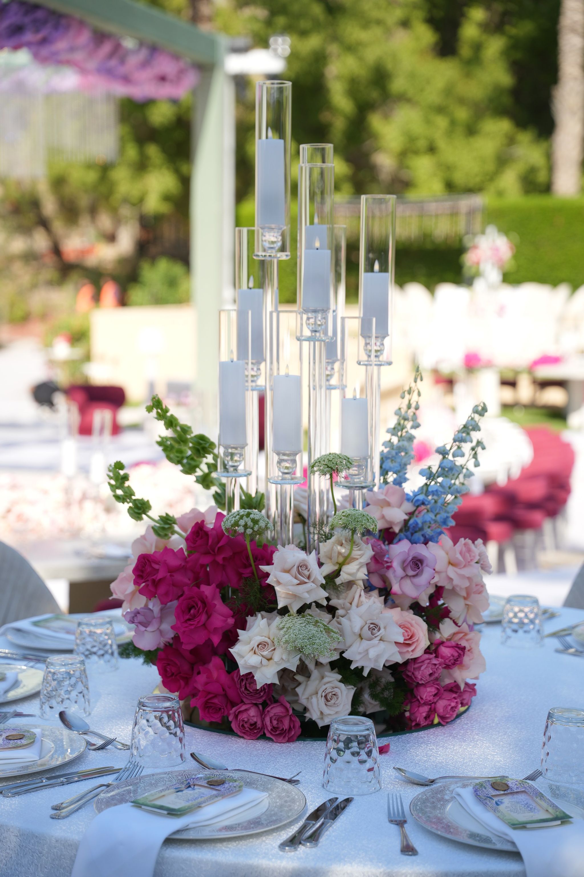 Glass pillar candles above a centrepiece of fuchsia roses, bells of Ireland and blue delphinium
