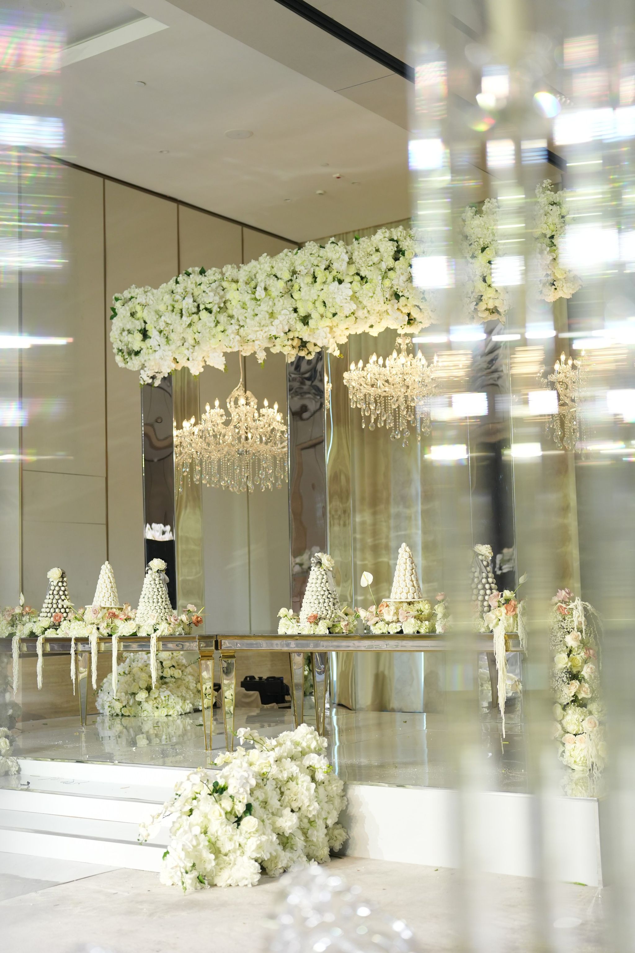Ivory hydrangea canopy and chandeliers above the mirrored stage, seen through soft crystal reflections