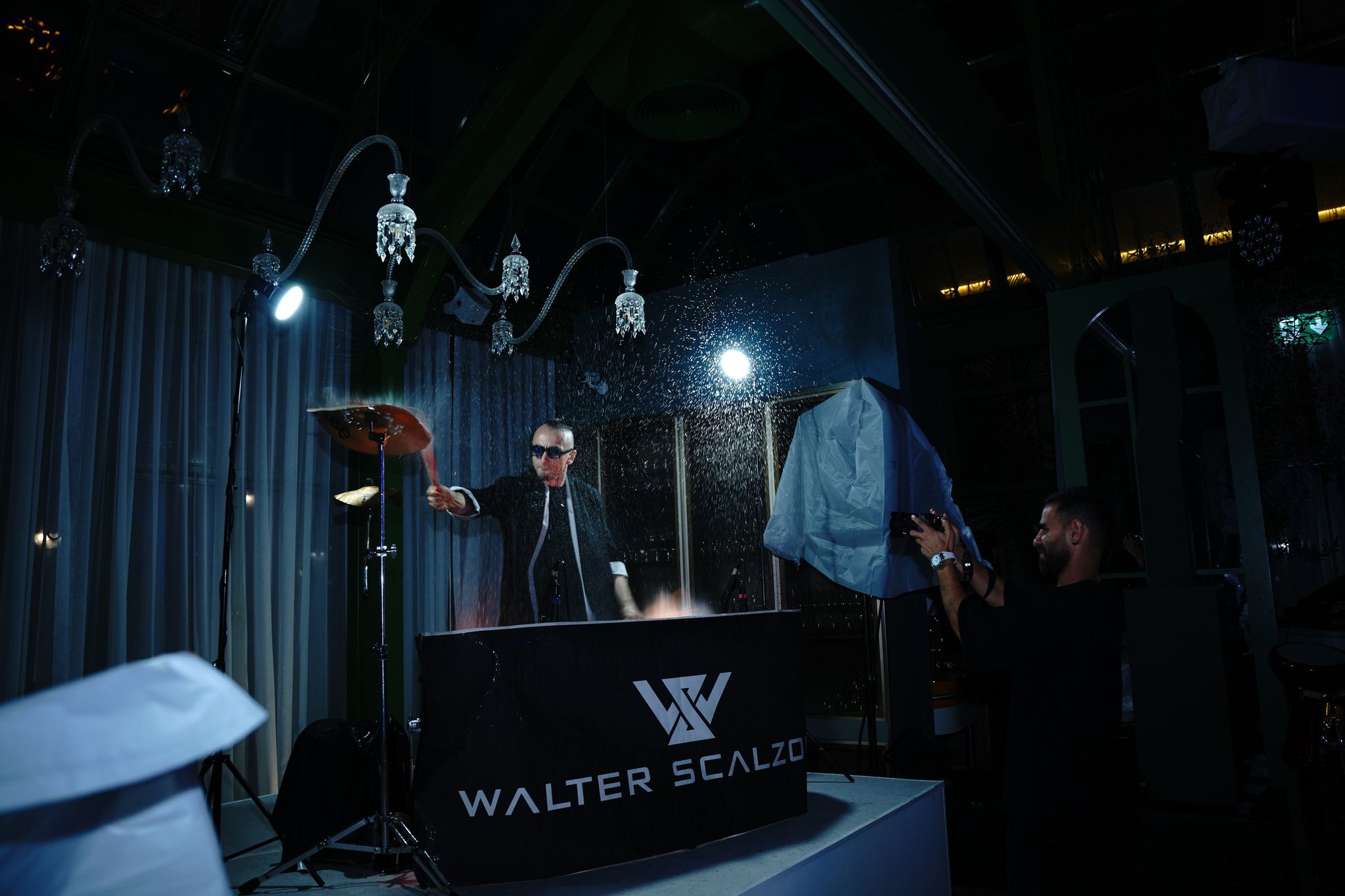 Percussionist at a branded DJ booth beneath crystal chandeliers as a guest films