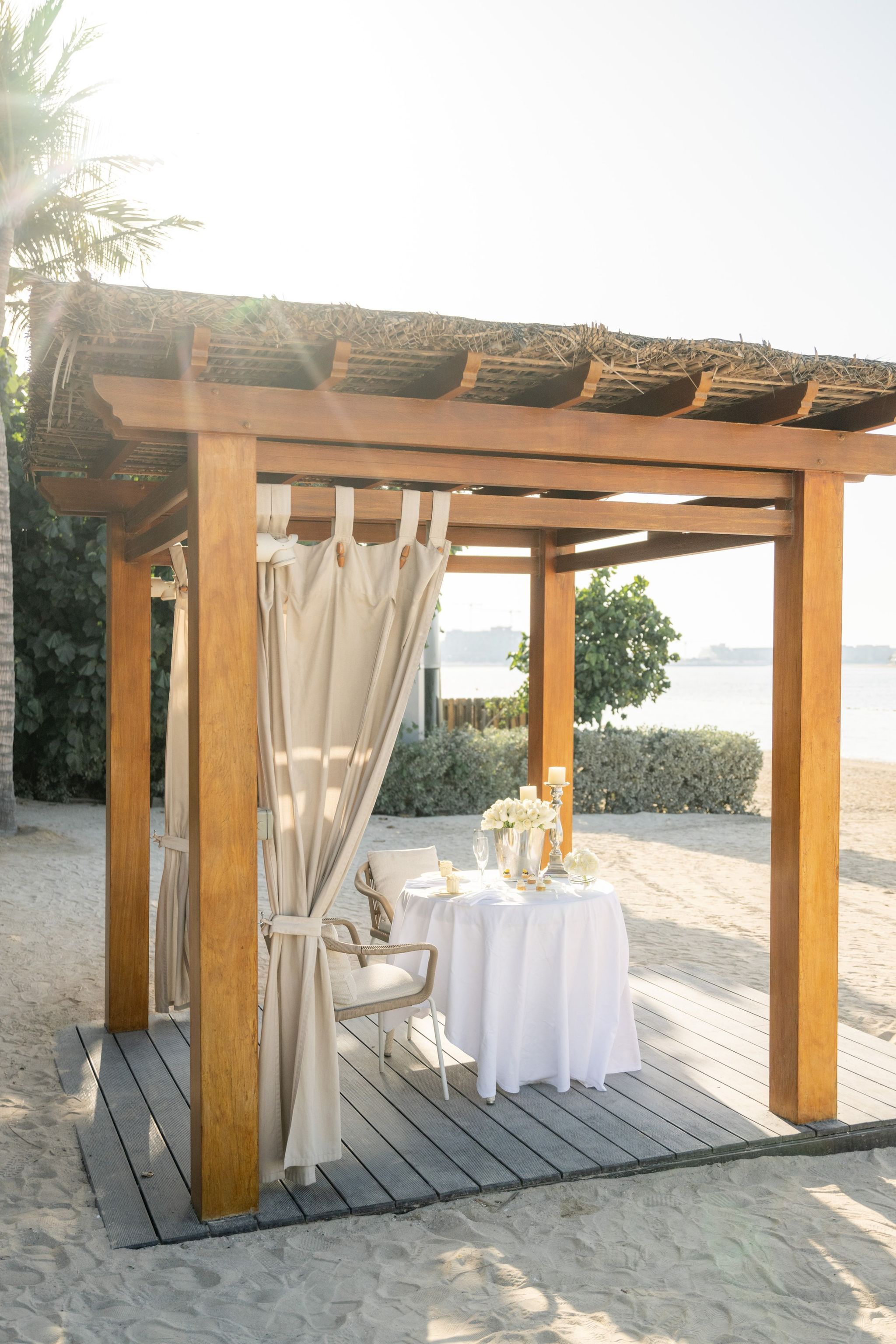 Timber beach cabana with linen drapes sheltering a white-clothed table for two
