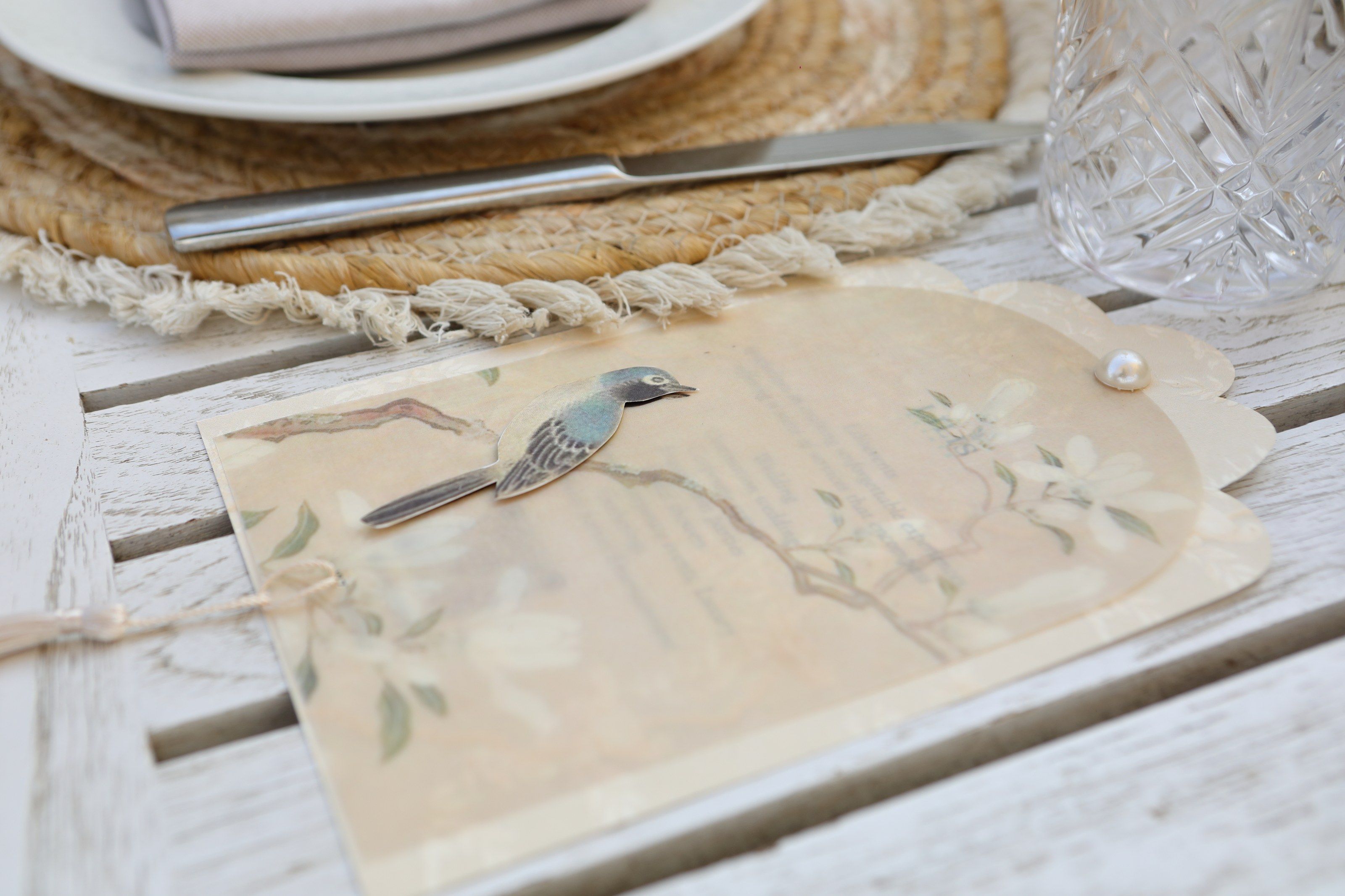 Illustrated menu card with a blue bird, magnolia branches and a pearl, set on a rattan placemat