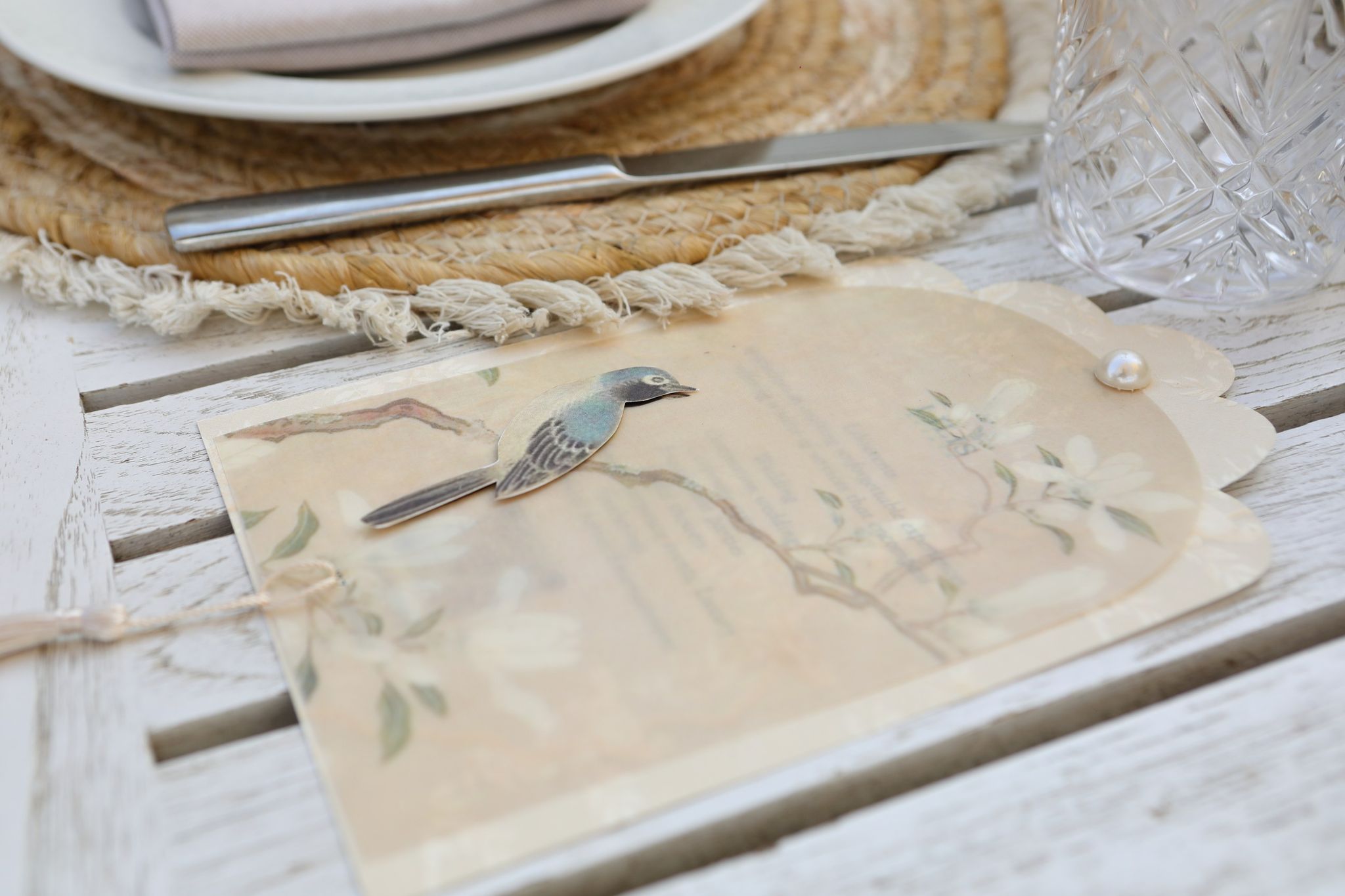 Illustrated menu card with a blue bird, magnolia branches and a pearl, set on a rattan placemat