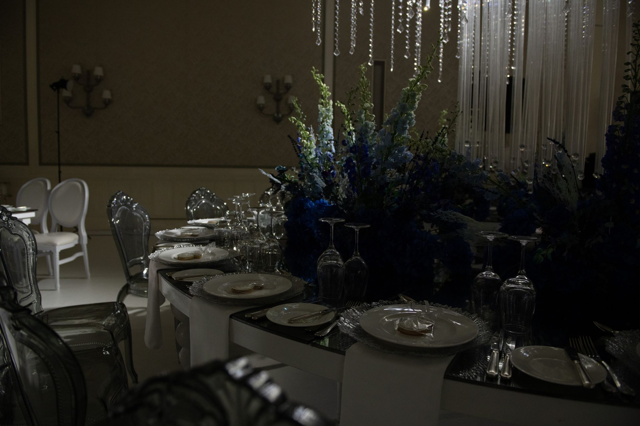 Table settings with plated favours and glassware beside tall blue florals in low light