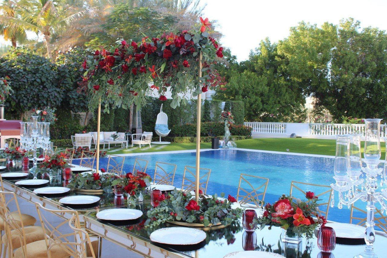 Long mirrored Yalda table with gold chairs and tall red floral stands beside a garden pool