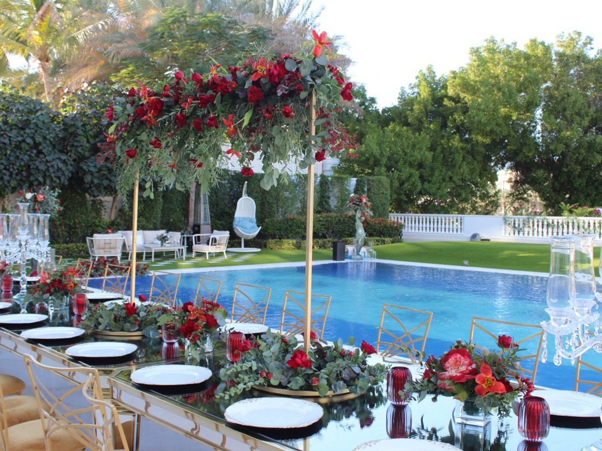 Long mirrored Yalda table with gold chairs and tall red floral stands beside a garden pool