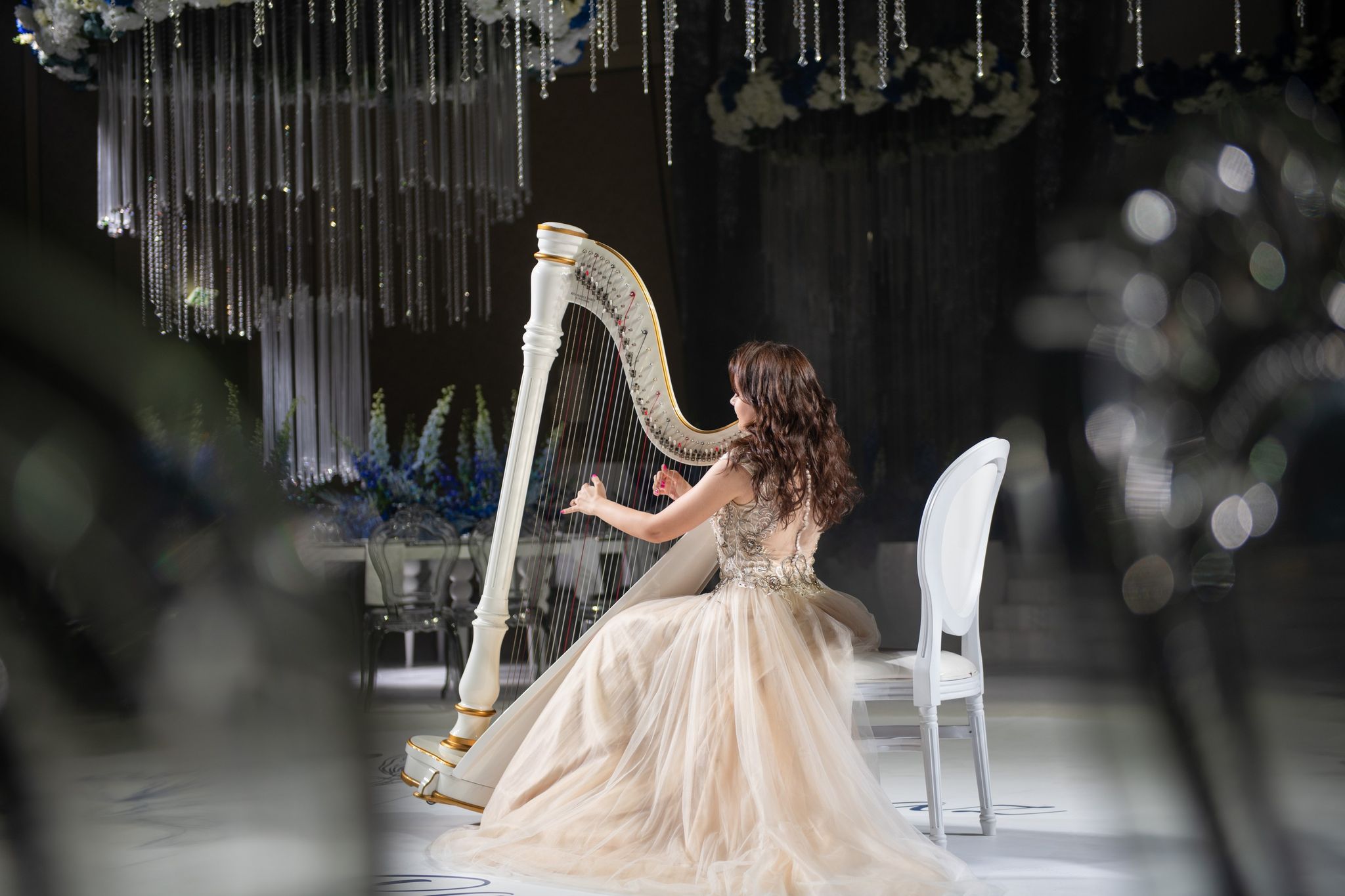 A harpist in a champagne gown playing a white and gold harp, crystal strands behind