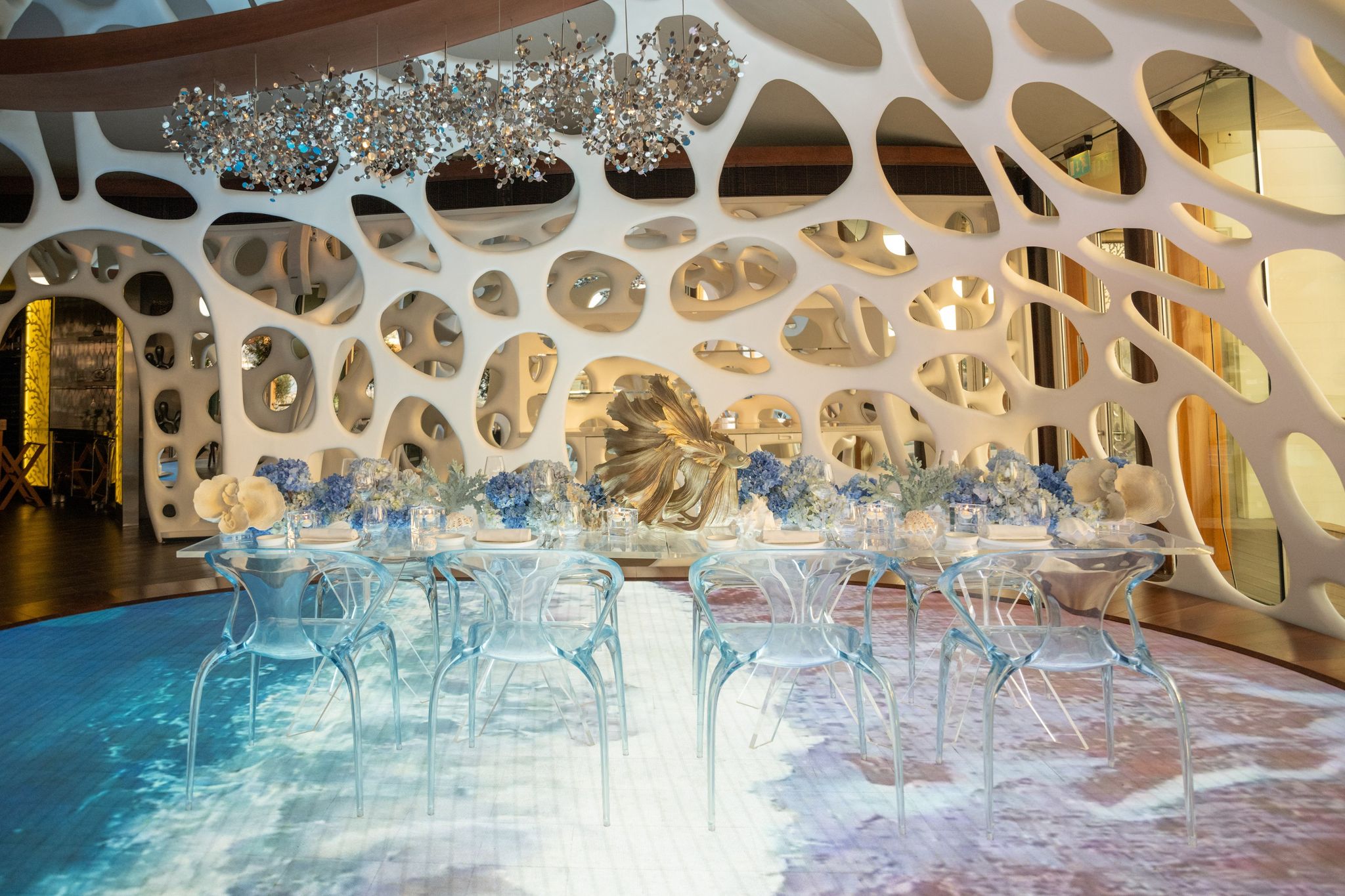 Dining room of sculpted white perforated walls, clear acrylic chairs, blue hydrangeas and a projected ocean floor