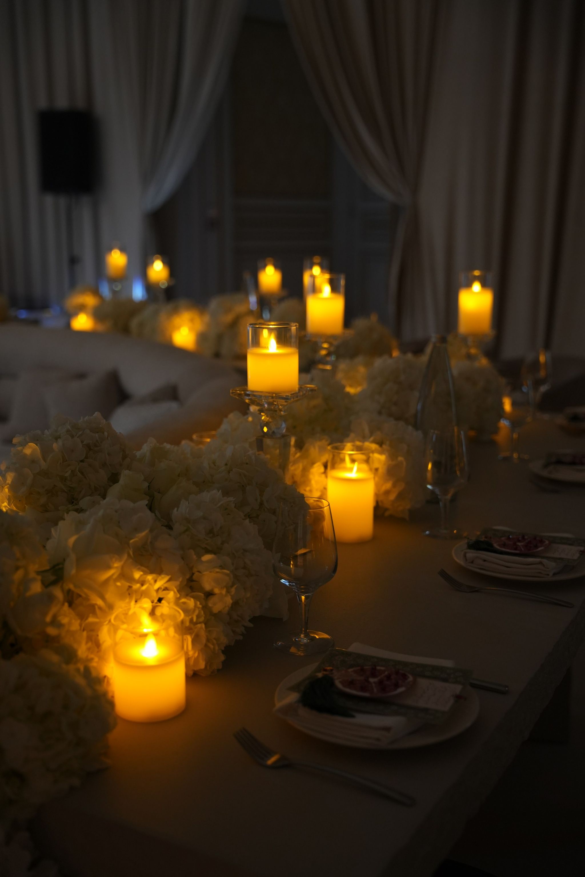 Candlelit table of white hydrangeas and glassware in a dimly lit room before guests arrive