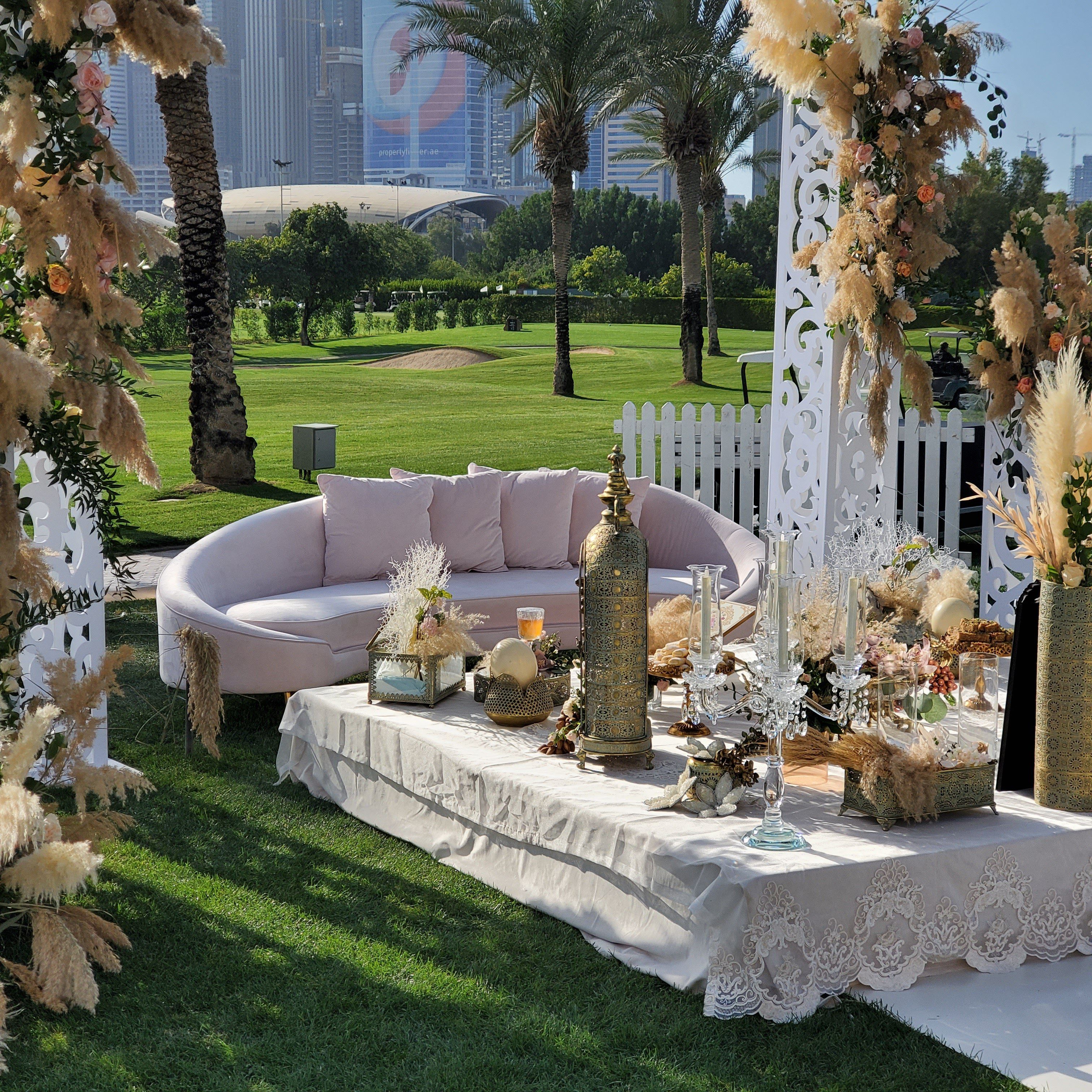 Blush curved sofa and lace-draped ceremony table framed by carved white panels and pampas at Emirates Golf Club