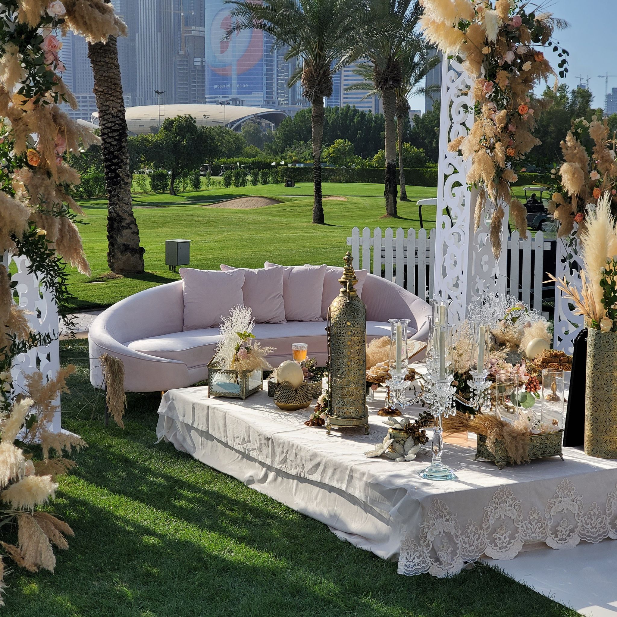 Blush curved sofa and lace-draped ceremony table framed by carved white panels and pampas at Emirates Golf Club