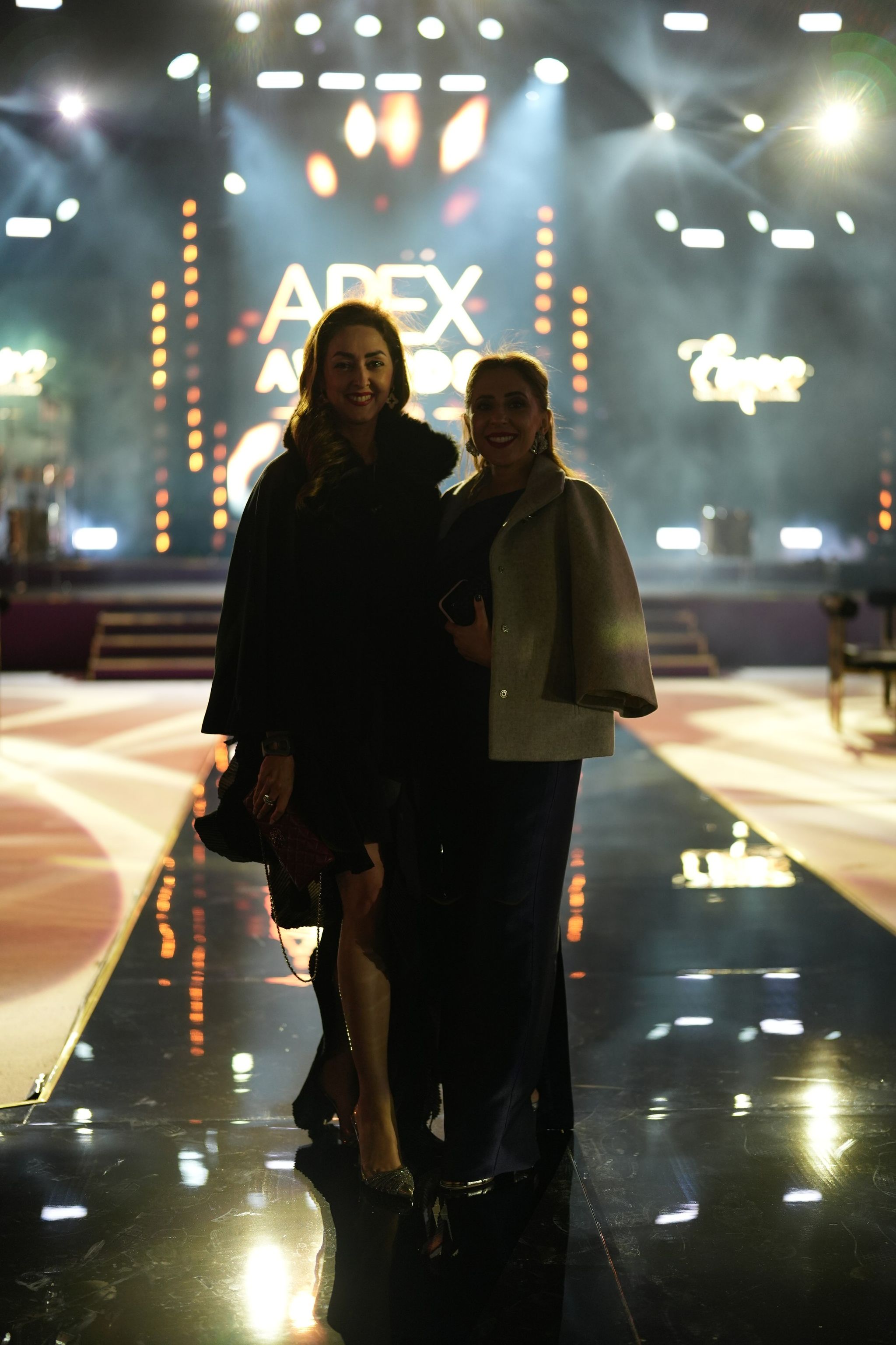 Two guests on a mirrored runway before an illuminated awards stage at an evening industry event