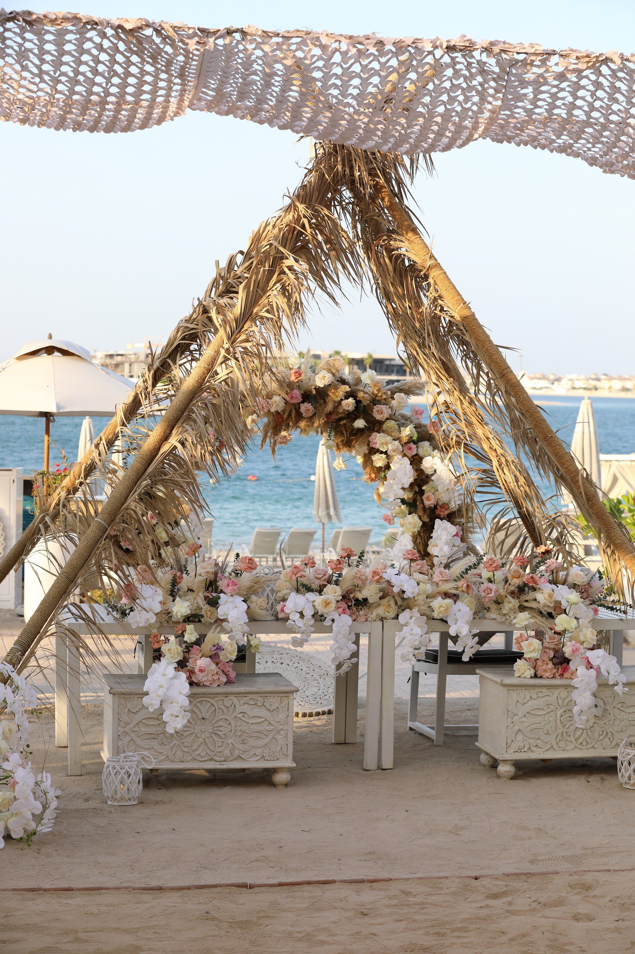 Palm-frond canopy dressed with blush roses and orchids above carved white tables on the beach at Jumeirah Zabeel Saray
