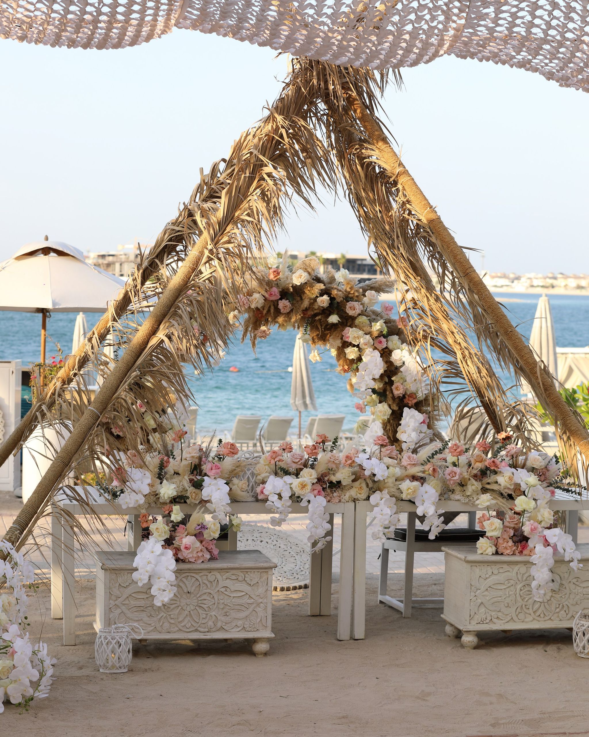 Palm-frond canopy dressed with blush roses and orchids above carved white tables on the beach at Jumeirah Zabeel Saray
