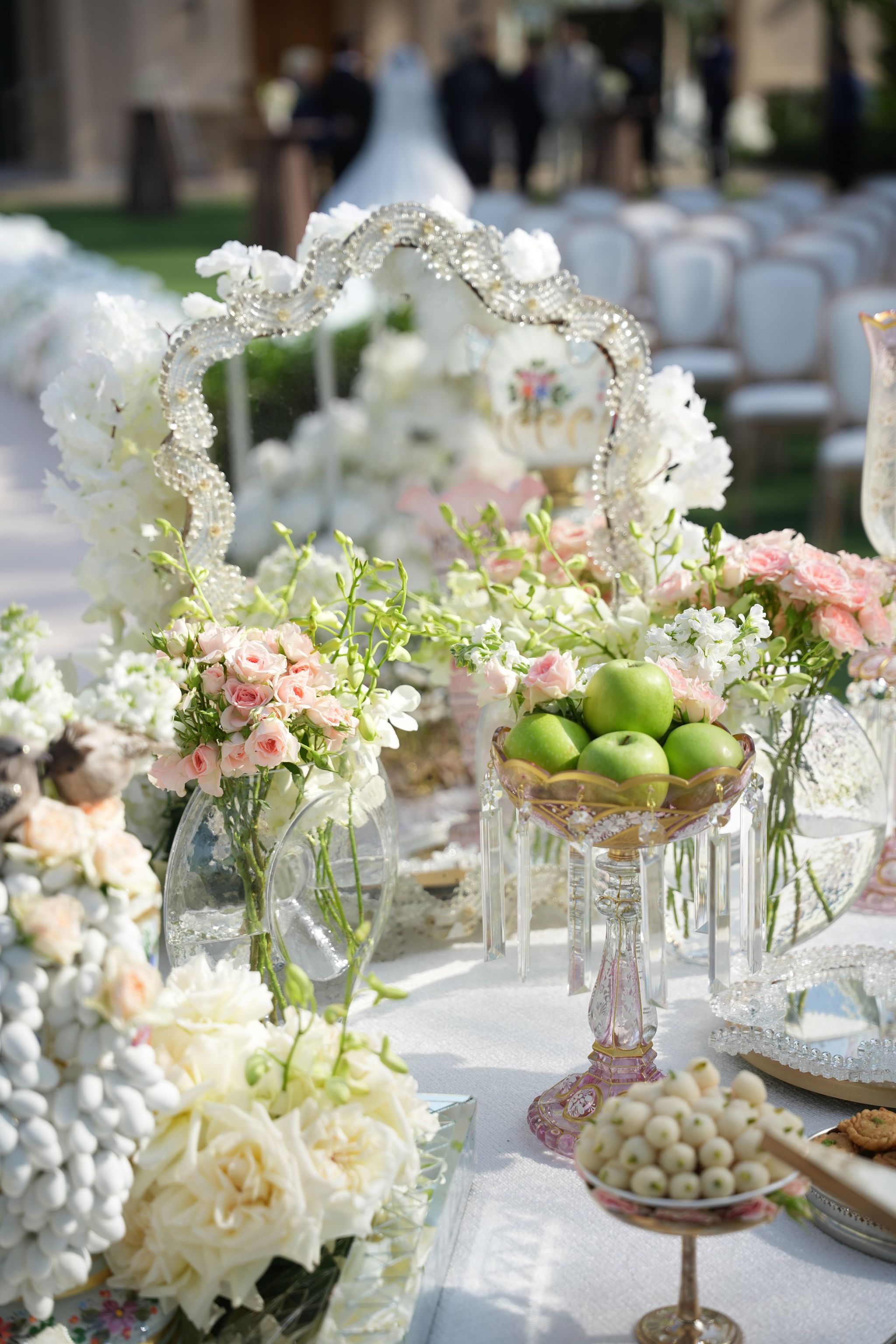Green apples in a gilded crystal compote beside a beaded mirror frame, pink roses and white orchids