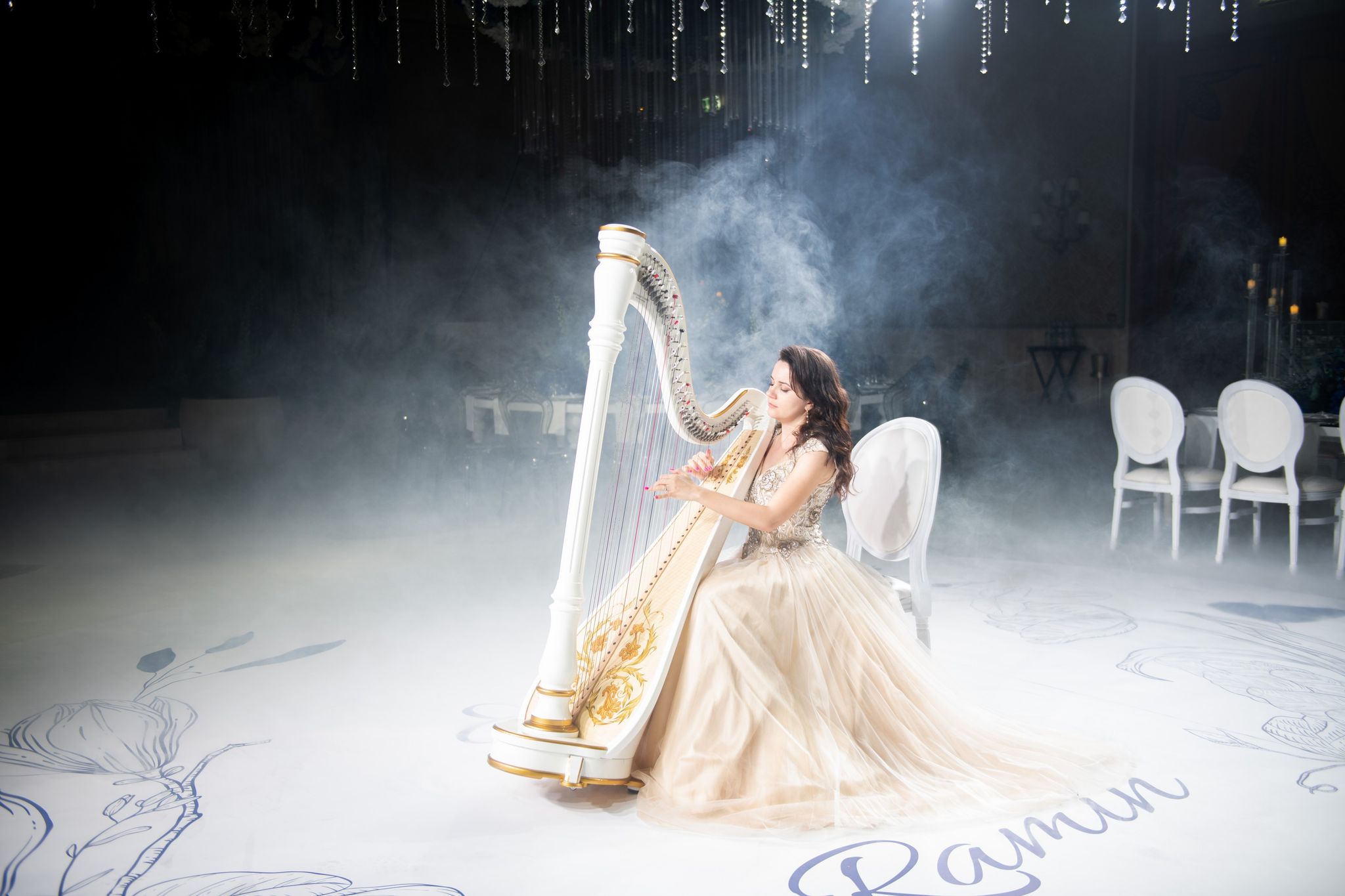 A harpist seated on a monogrammed dance floor as haze drifts under crystal strands