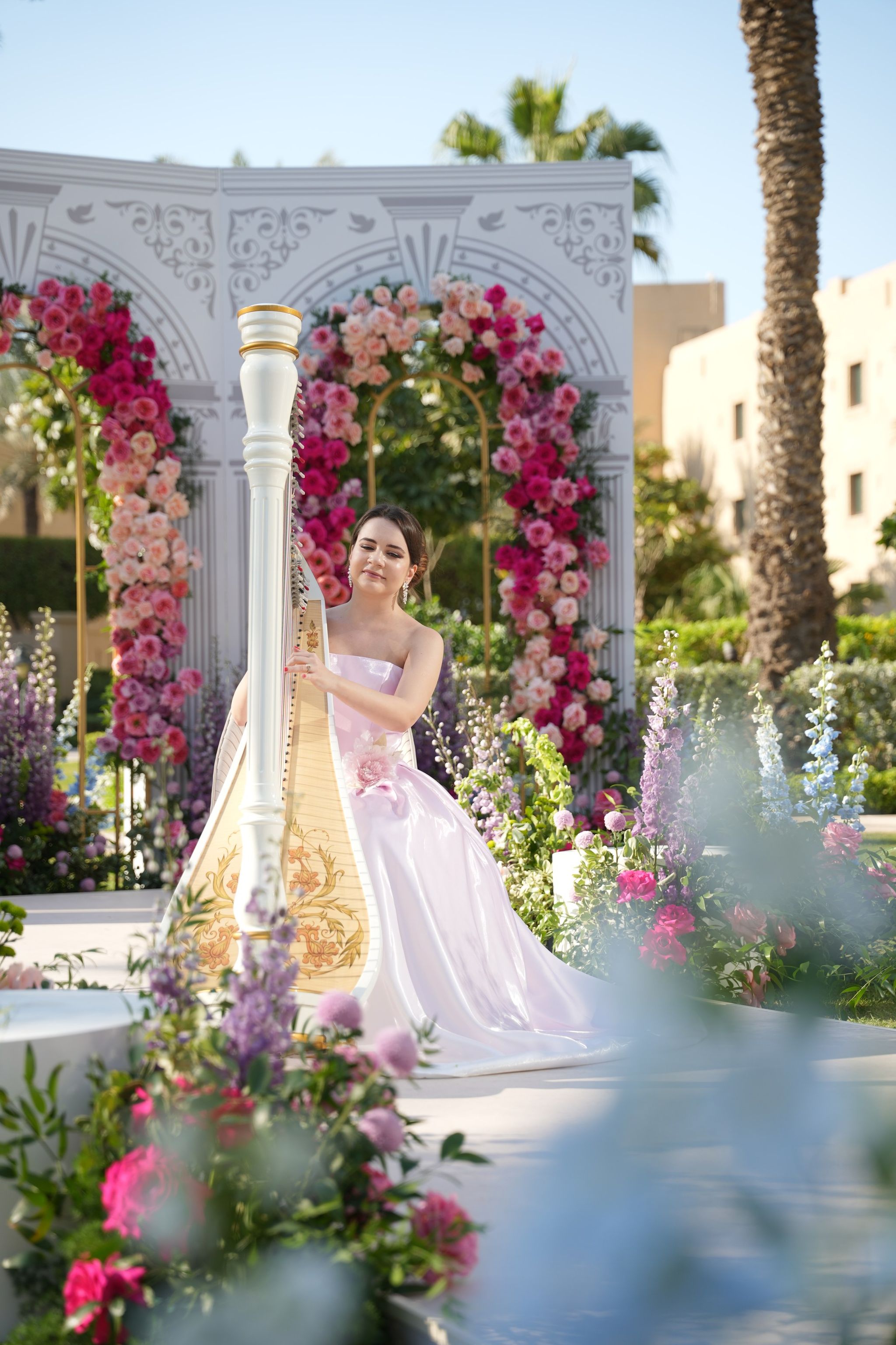 Harpist playing a gilded white harp beside delphiniums and pink roses on the aisle