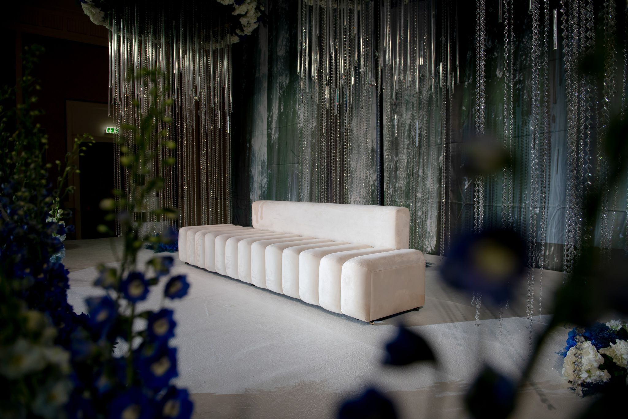 Channel-tufted ivory sofa before curtains of crystal, framed by out-of-focus blue delphiniums
