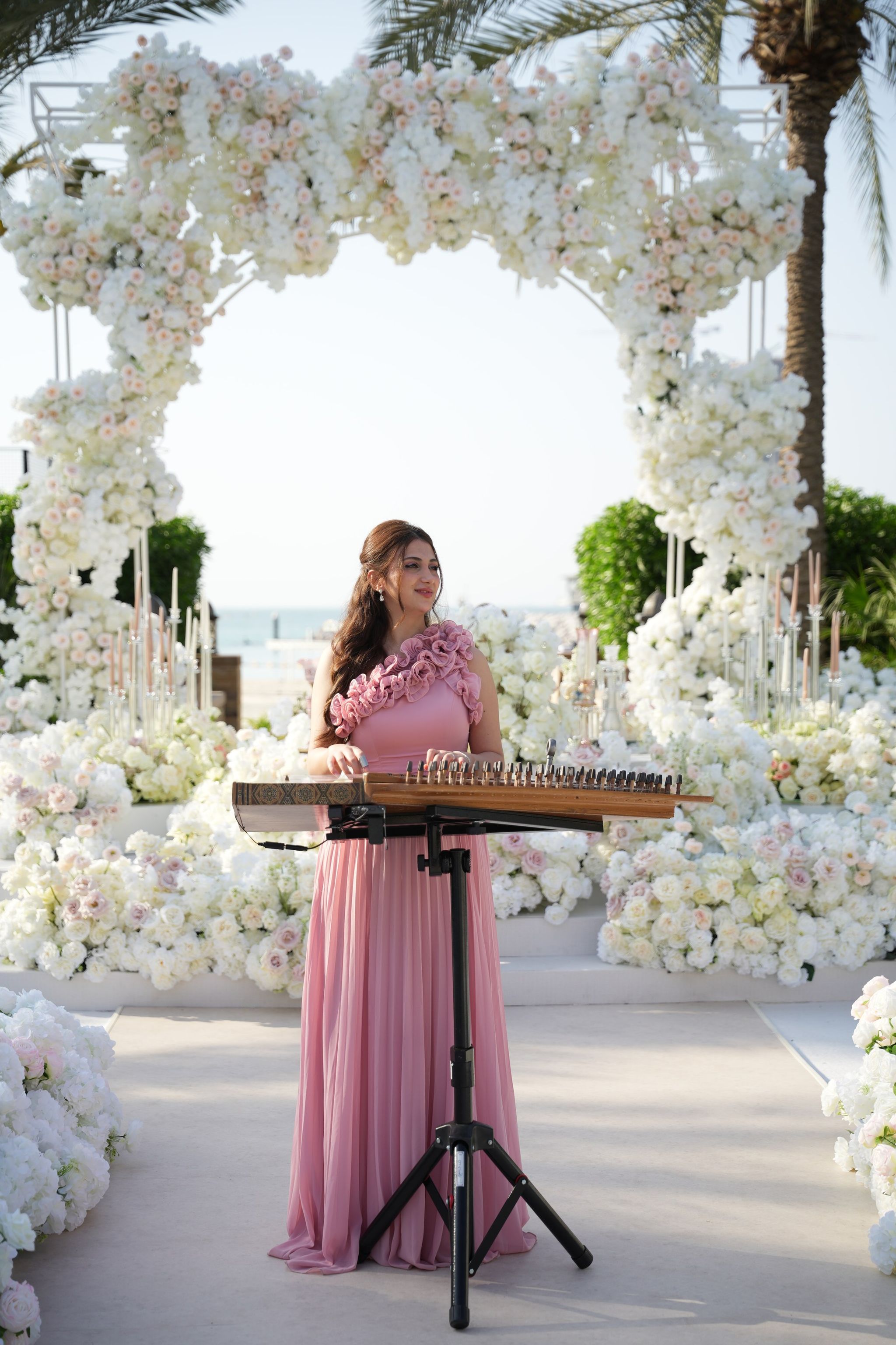 Qanun player in a blush gown performing before the floral arch, with the sea beyond