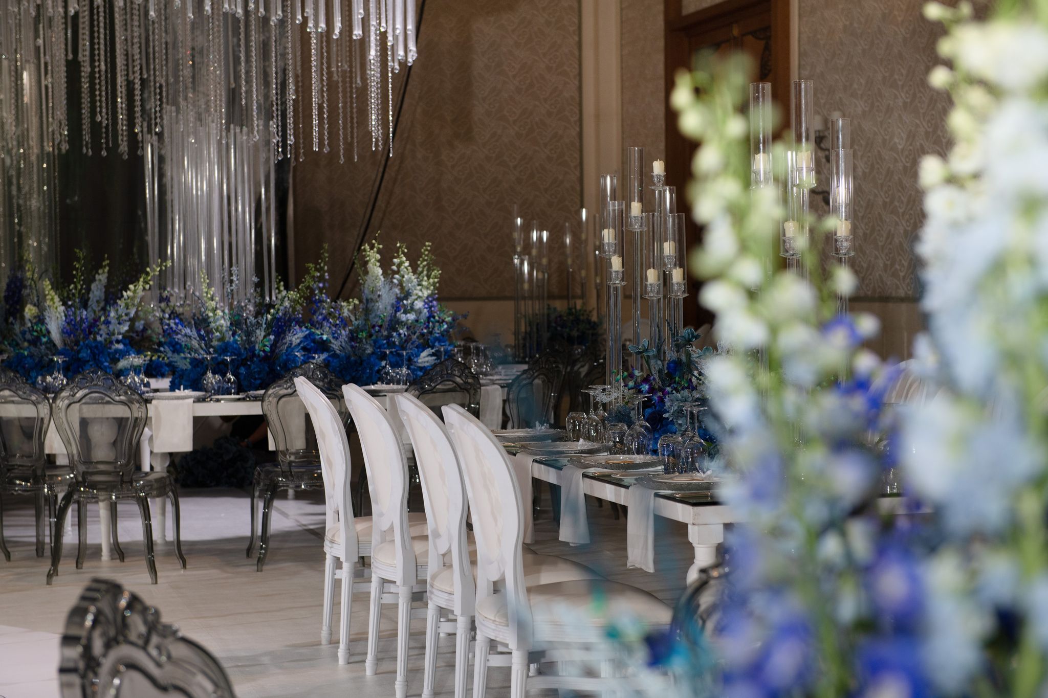 White chairs at a candlelit long table, with blue florals and crystal strands beyond