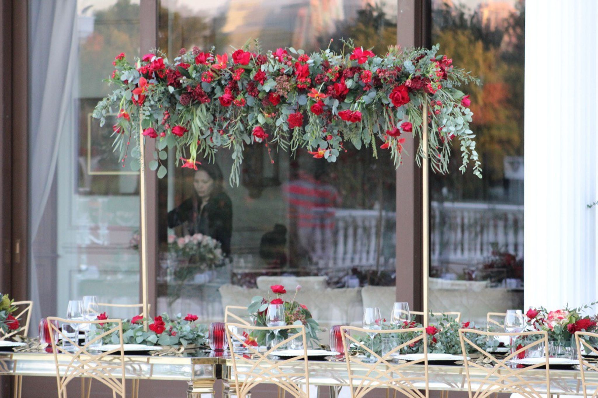 Suspended garland of red roses and eucalyptus above a mirrored table set before glass doors