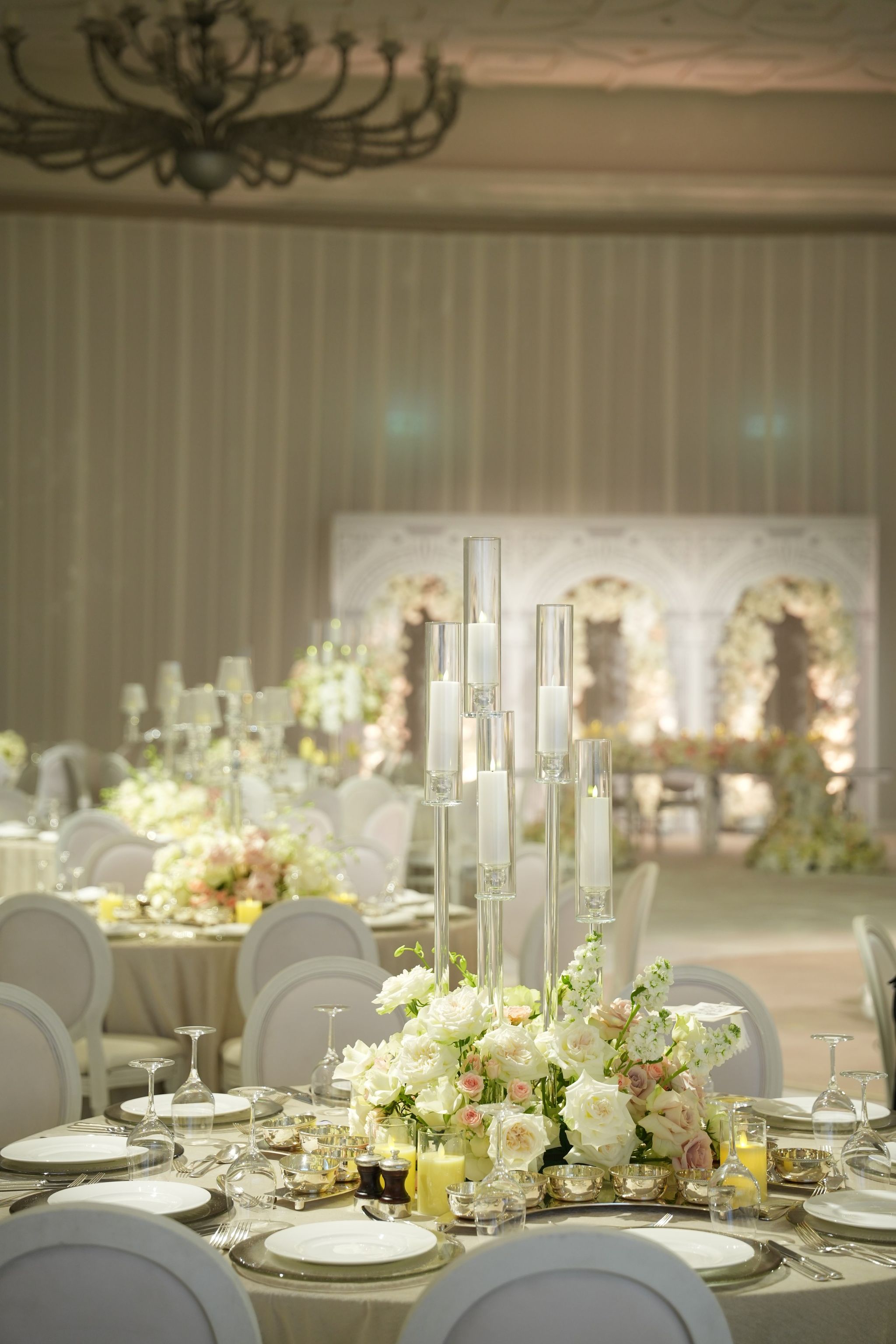 Glass-cylinder candelabra above low white and blush roses, a chandelier and floral arches in the distance