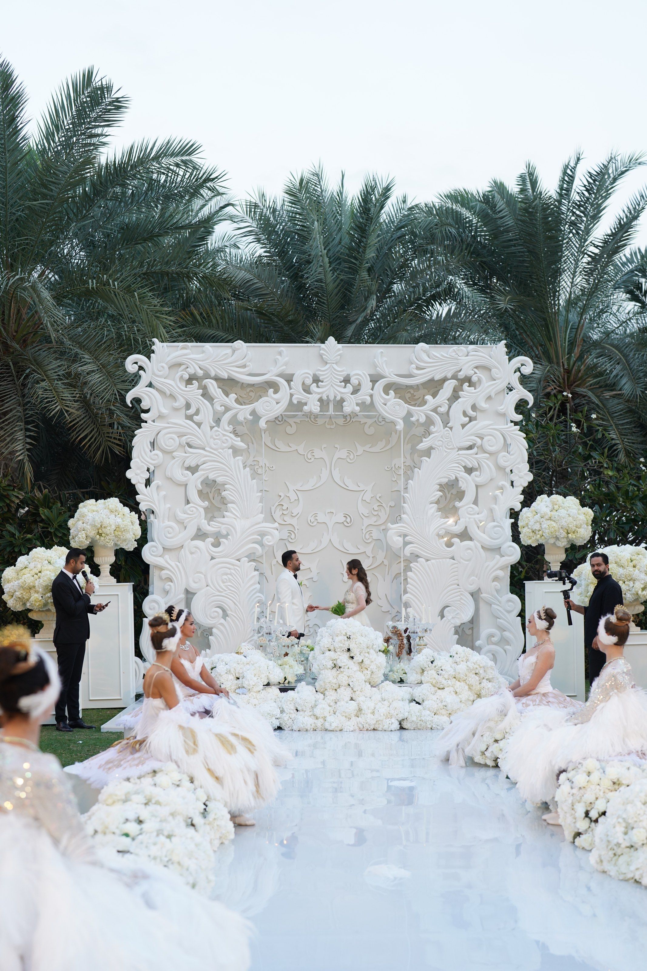The couple before the carved white backdrop as ballerinas in feathered tutus kneel along the aisle