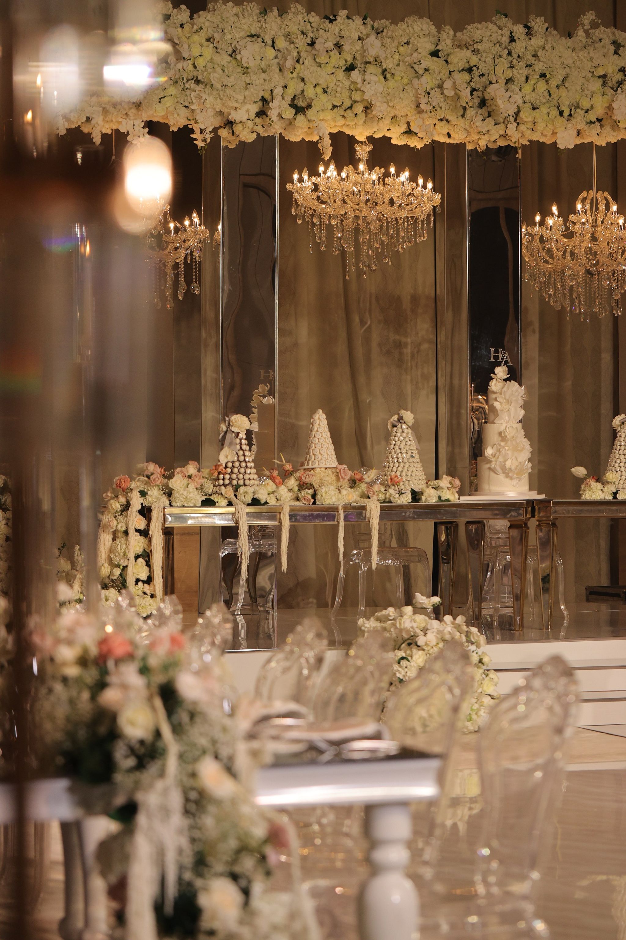 Tiered wedding cake and croquembouche towers on a mirrored table under crystal chandeliers