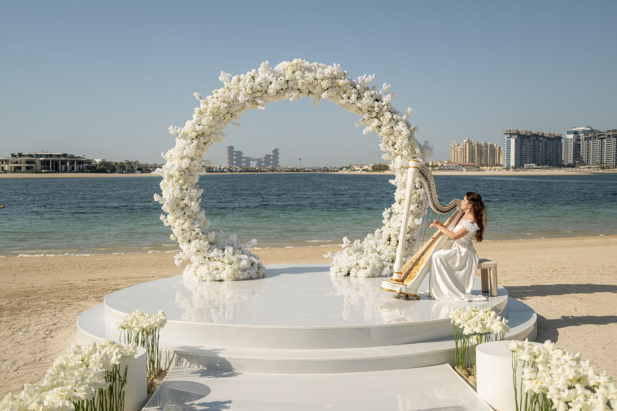 Circular arch of white blooms on a mirrored beach stage, a harpist playing beside it on Palm Jumeirah