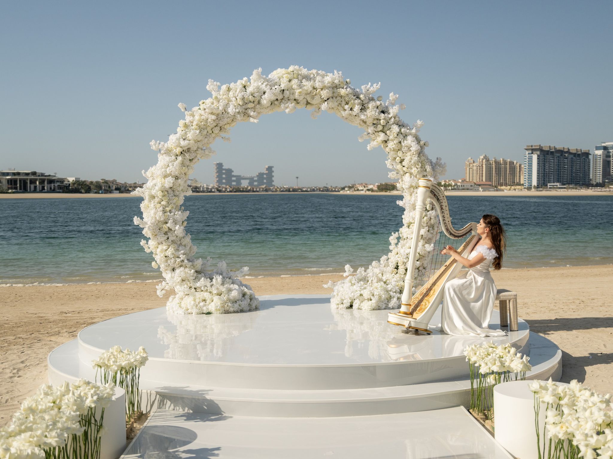 Circular arch of white blooms on a mirrored beach stage, a harpist playing beside it on Palm Jumeirah