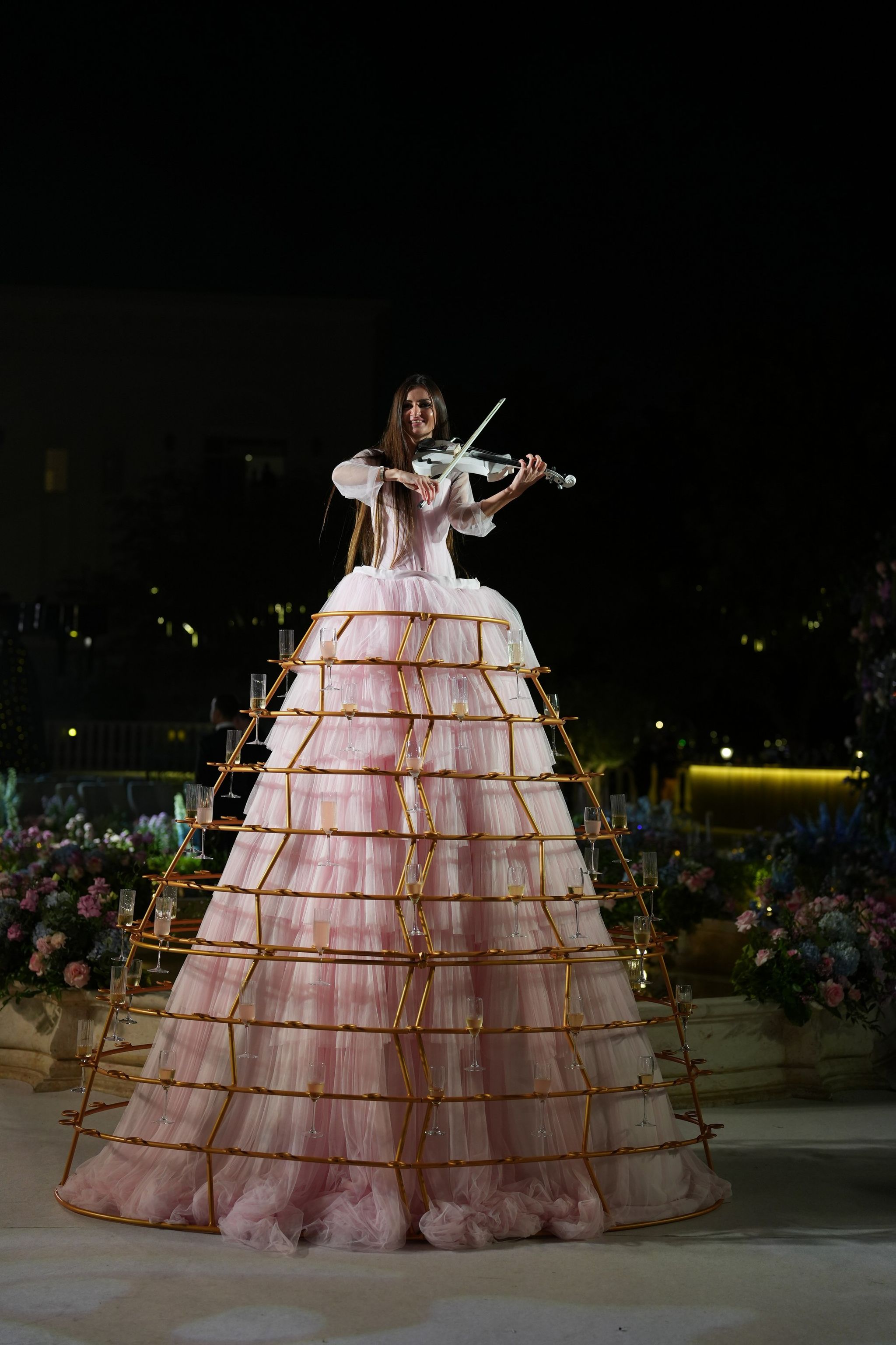 Violinist in a pink tulle gown inside a gilded frame holding champagne glasses at night
