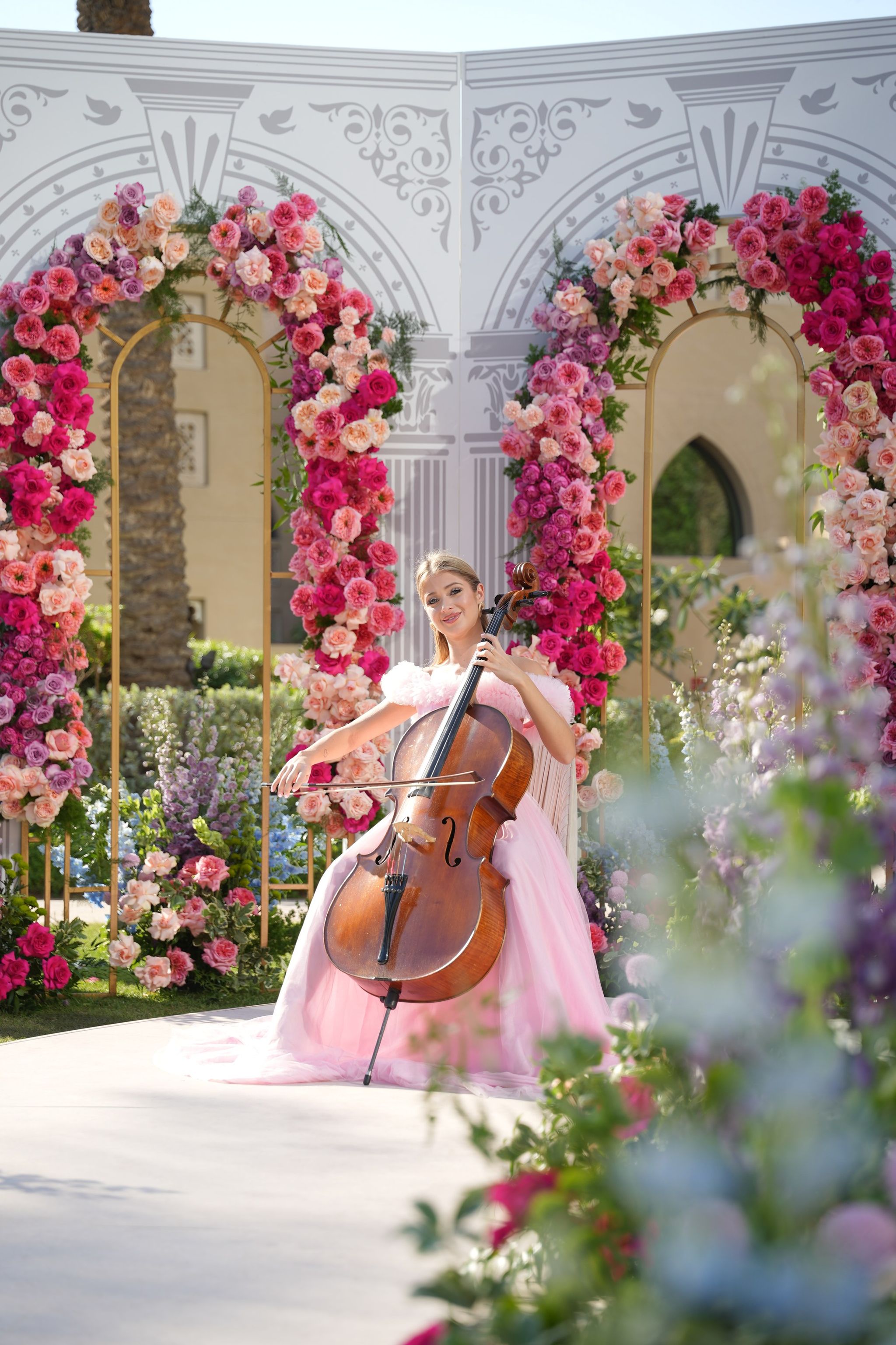 Cellist seated under the rose arches, framed by soft-focus delphiniums in the foreground