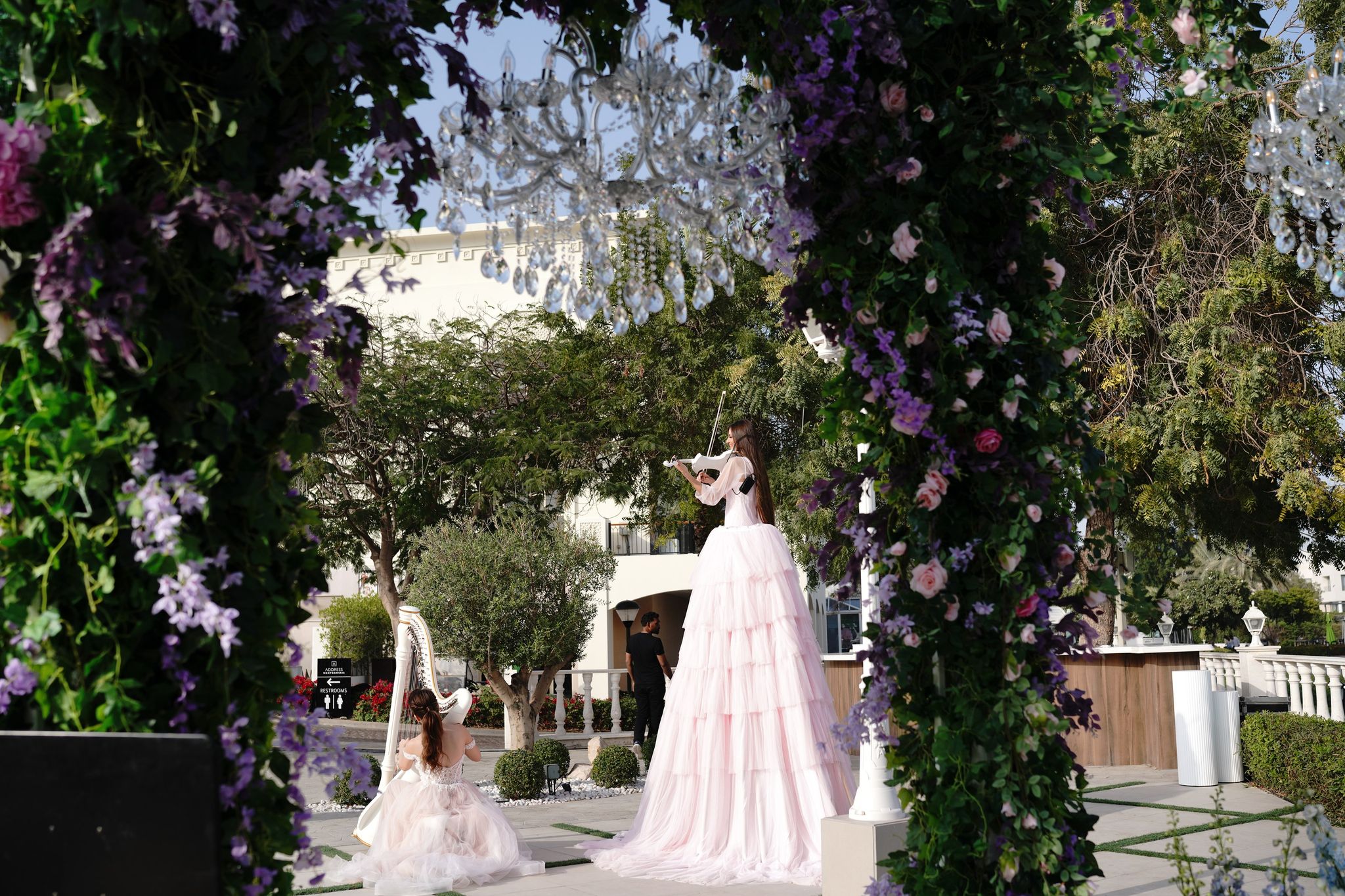 Stilt violinist and harpist performing beneath a wisteria arch hung with crystal chandeliers