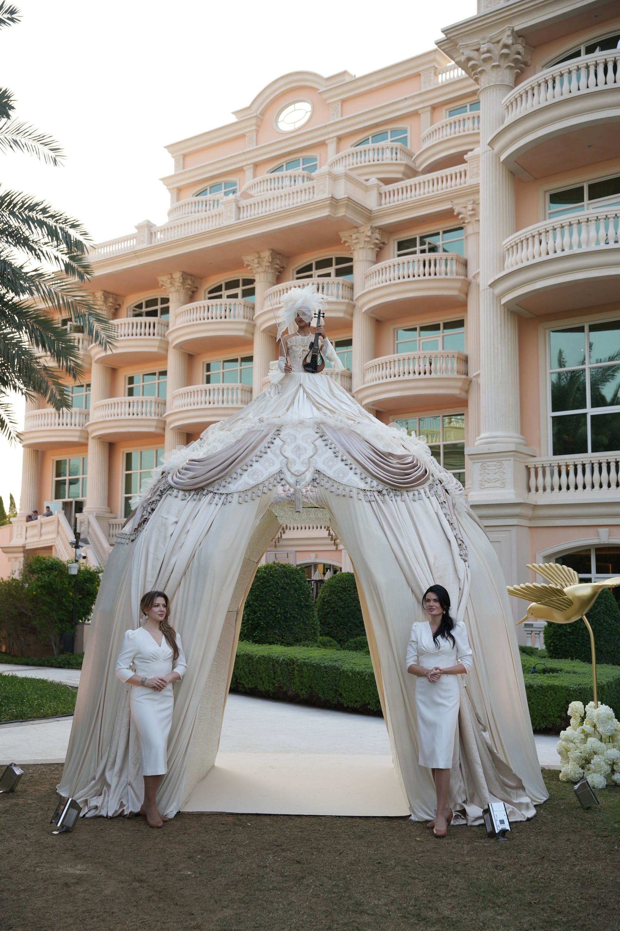 Draped ivory tent topped by a violinist in a feathered gown before the pink facade of Raffles The Palm Dubai