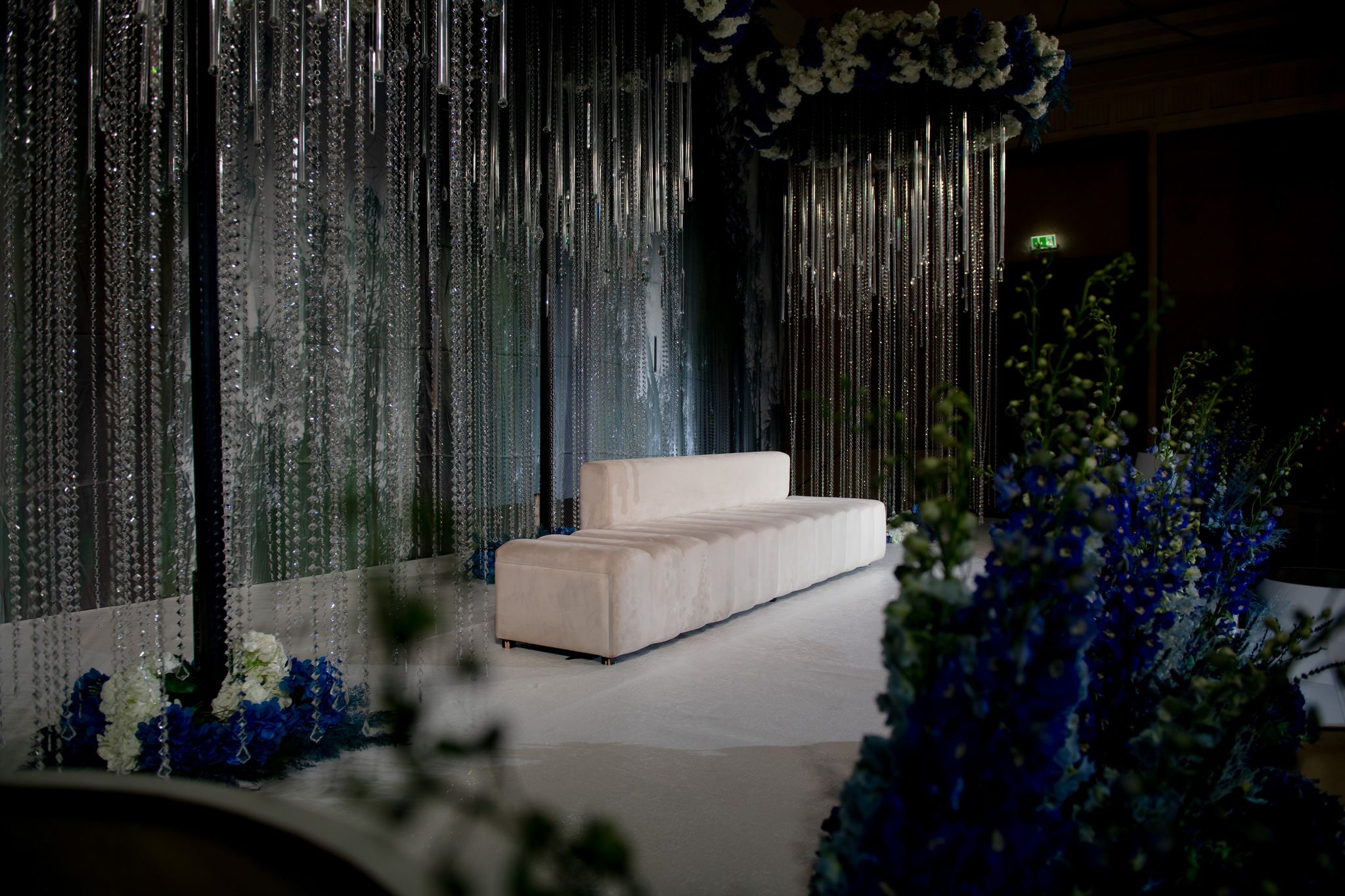 Ivory stage sofa between crystal curtains, with blue delphiniums and white roses in the foreground